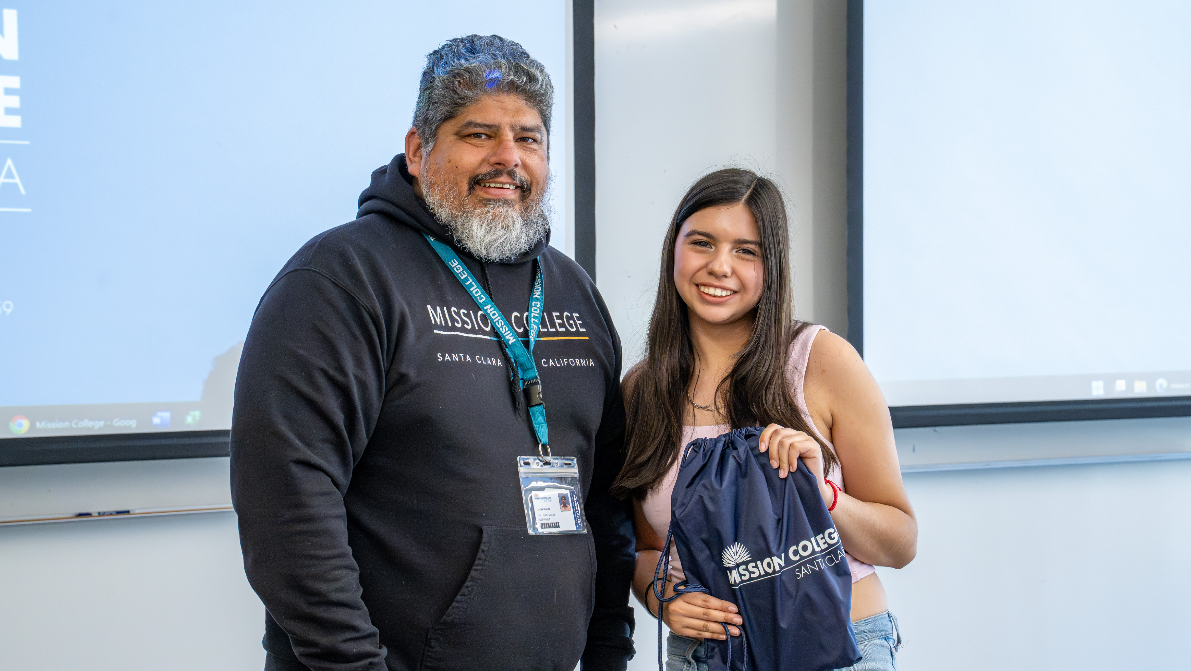 Mission College staff member in a black hoodie posing for a photo with a smiling student holding a blue Mission College cinch bag.