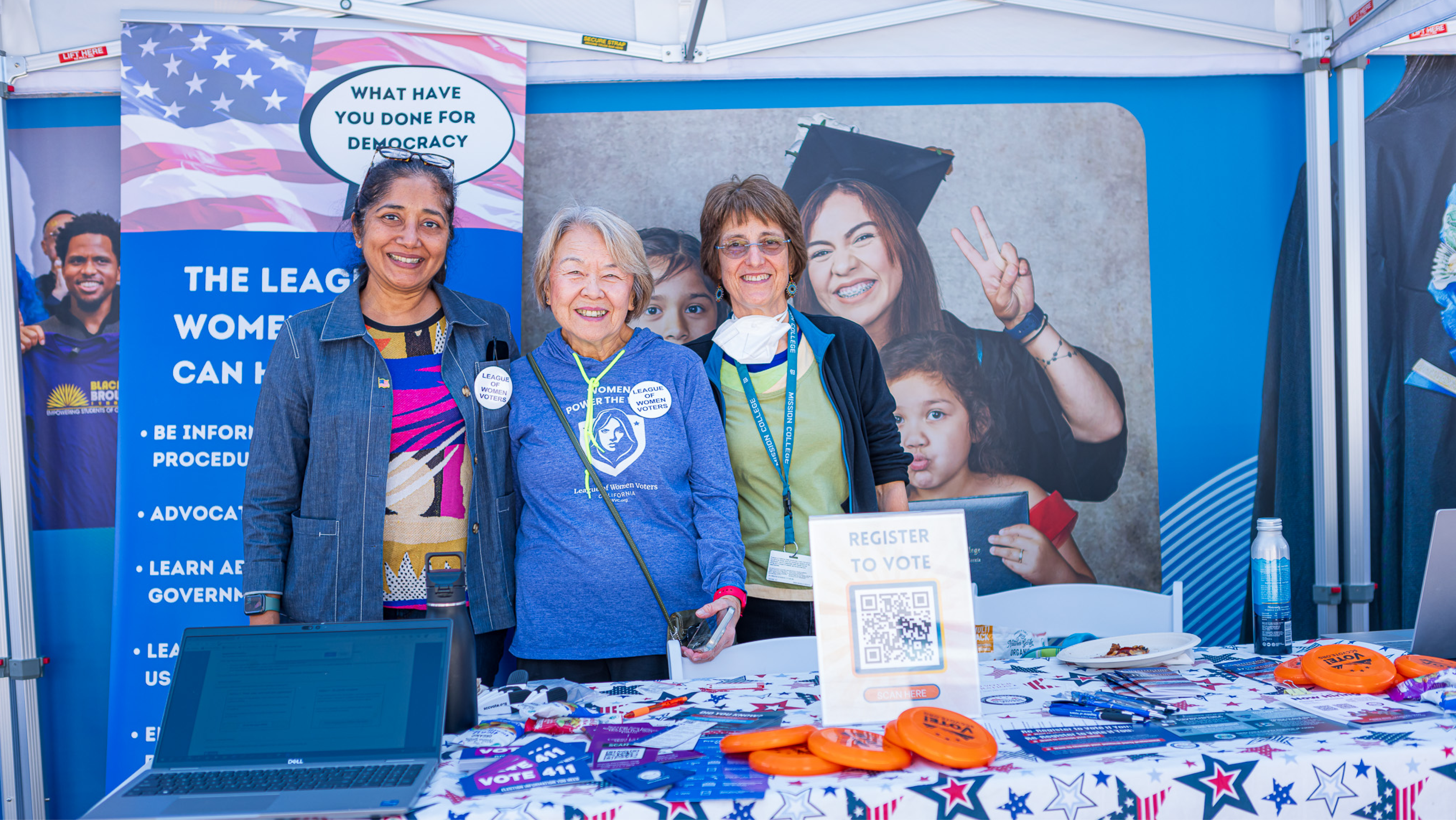 Three women posing together in front of a League of Women Voters booth with voter registration materials, laptops, and a QR code sign.