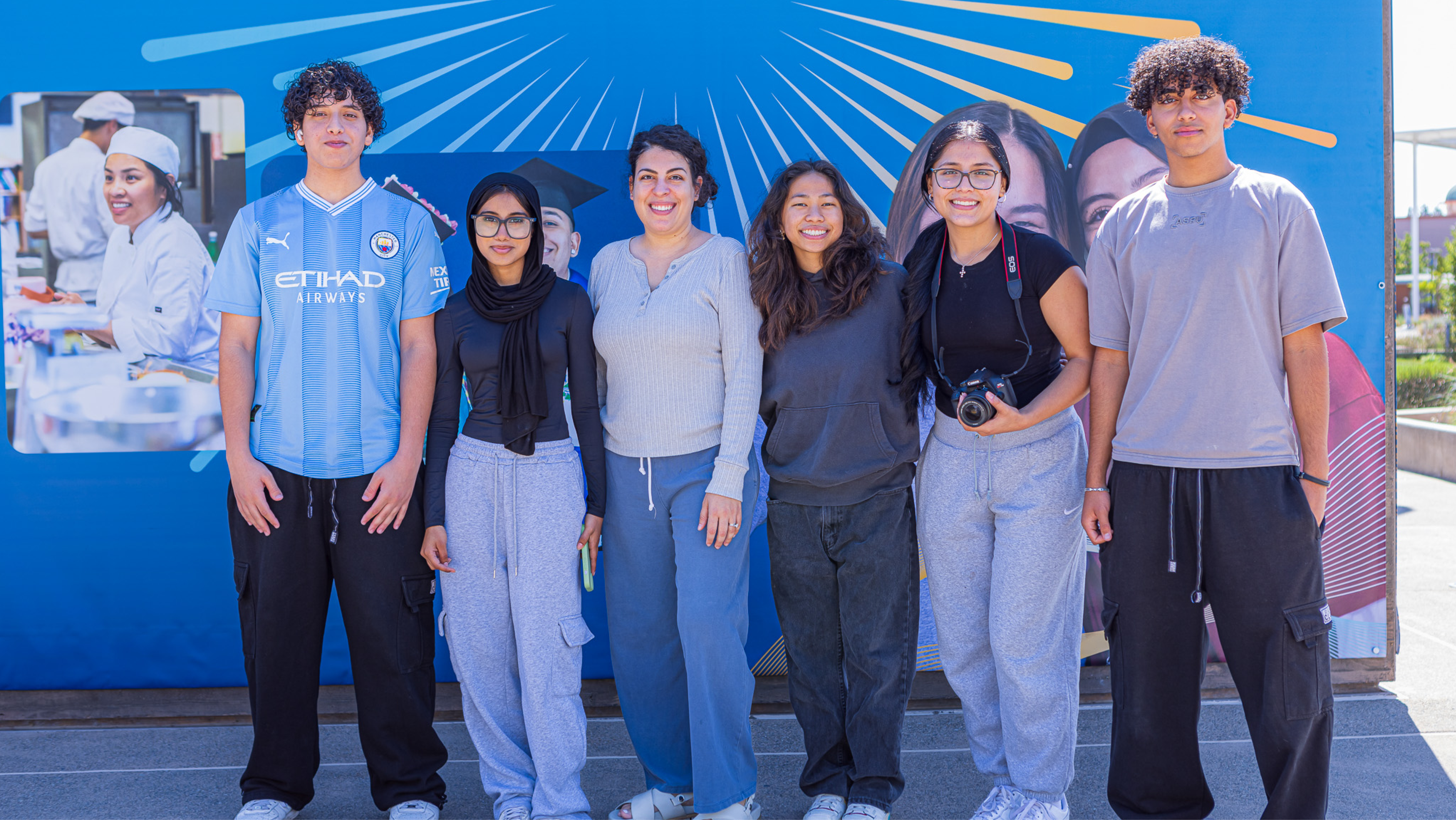 Group of six students smiling and posing together in a line in front of a blue campus backdrop outdoors.
