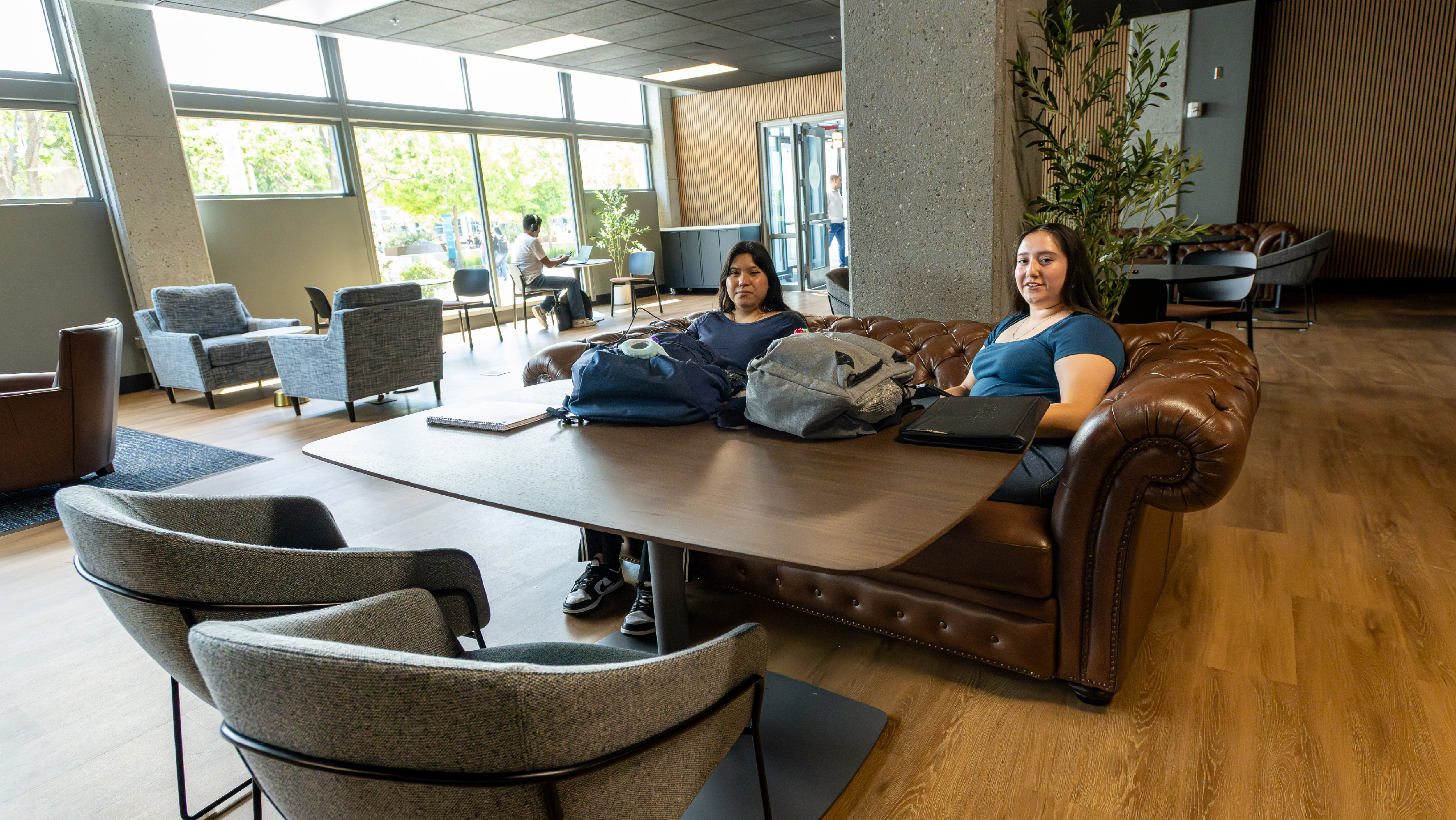 Two female students sitting at a large wooden conference table in a modern lounge area with comfortable leather seating.