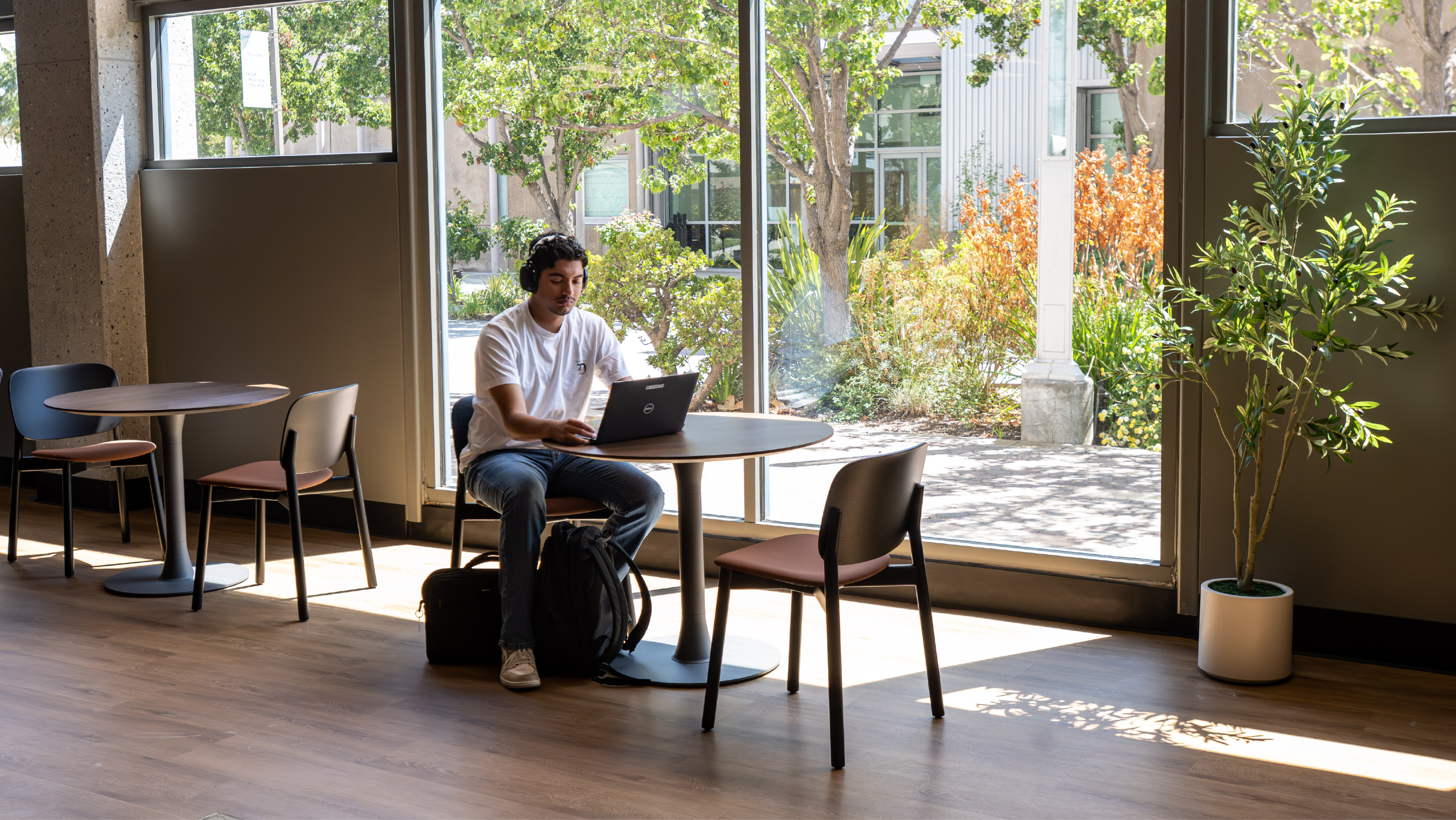 Male student wearing headphones working on a laptop at a round table near a large floor-to-ceiling window overlooking campus grounds.