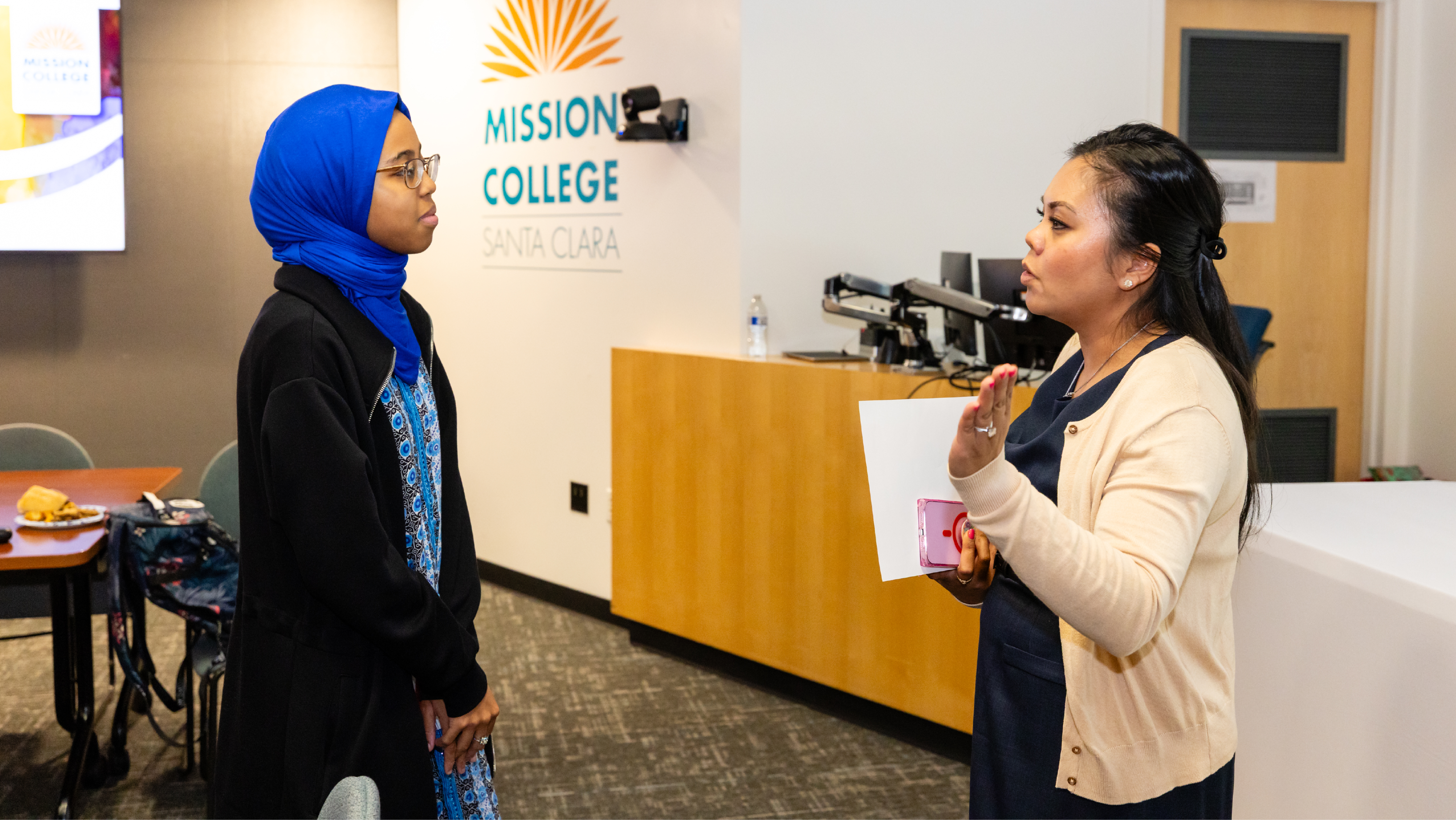 Faculty member speaking with a student wearing a blue hijab in an office setting in front of a Mission College logo.