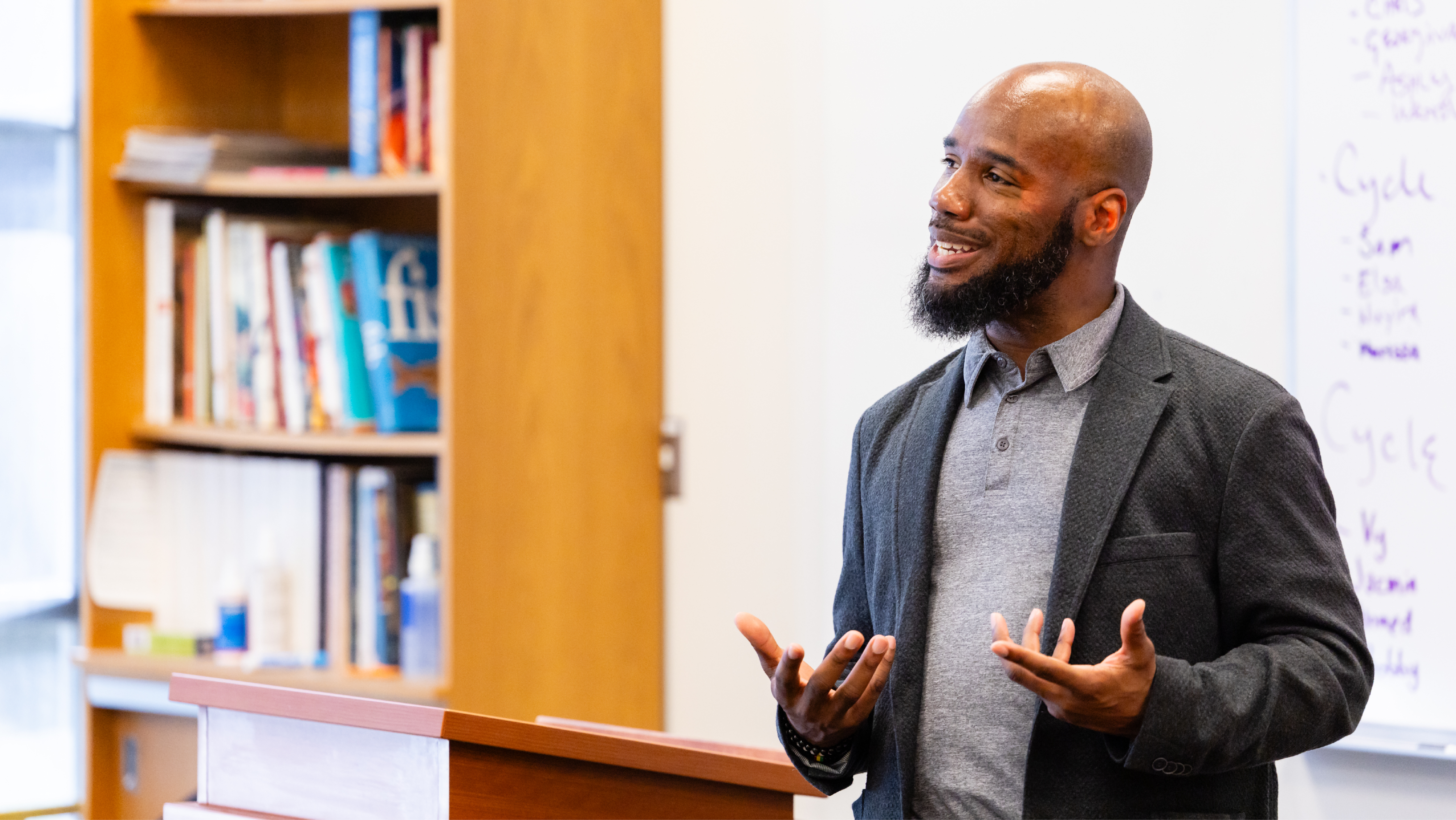 Presenter speaking at a wooden podium in a classroom with a whiteboard and bookshelf in the background.