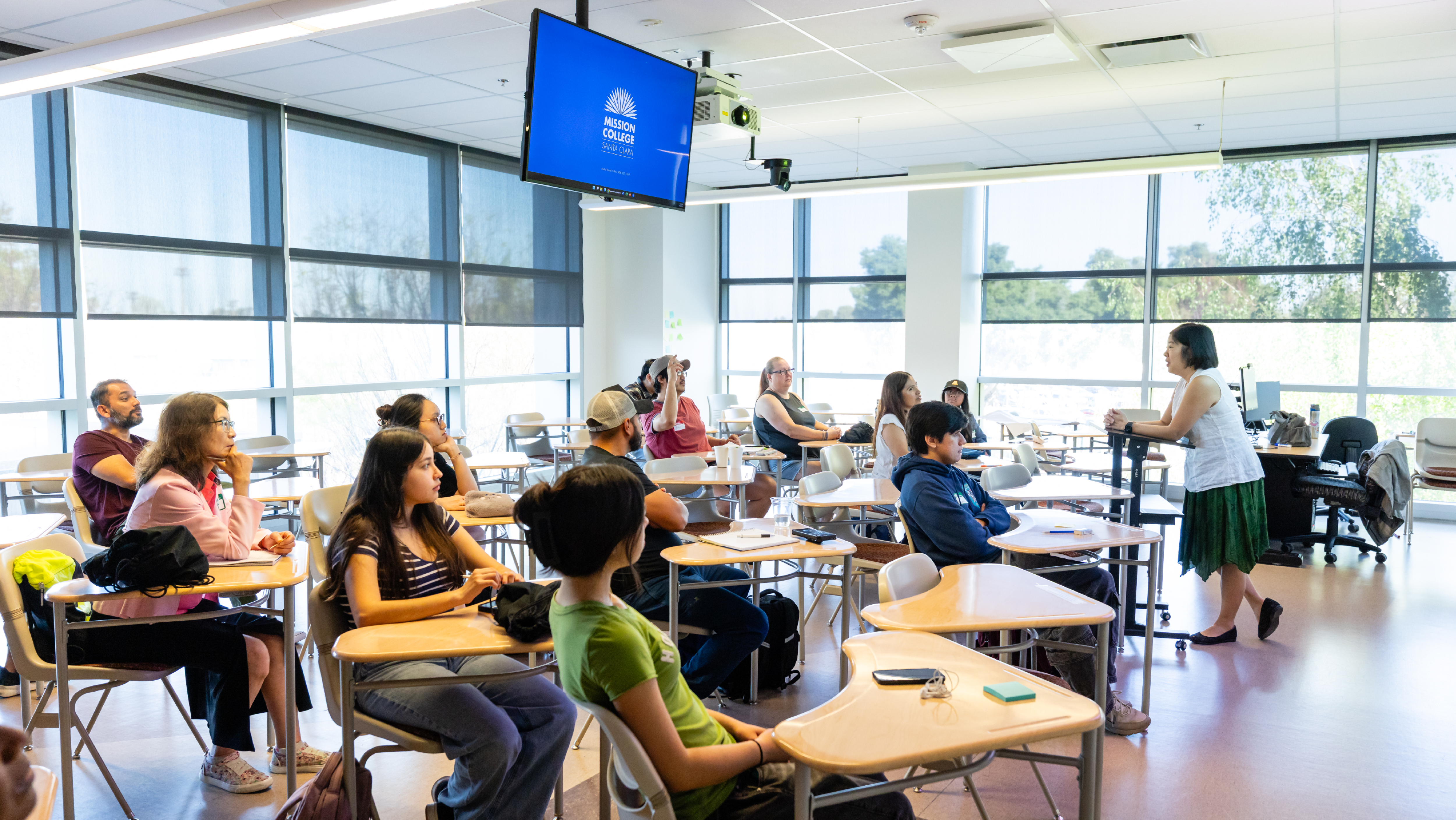 Instructor speaking to a classroom of attentive students seated at individual desks facing the front.