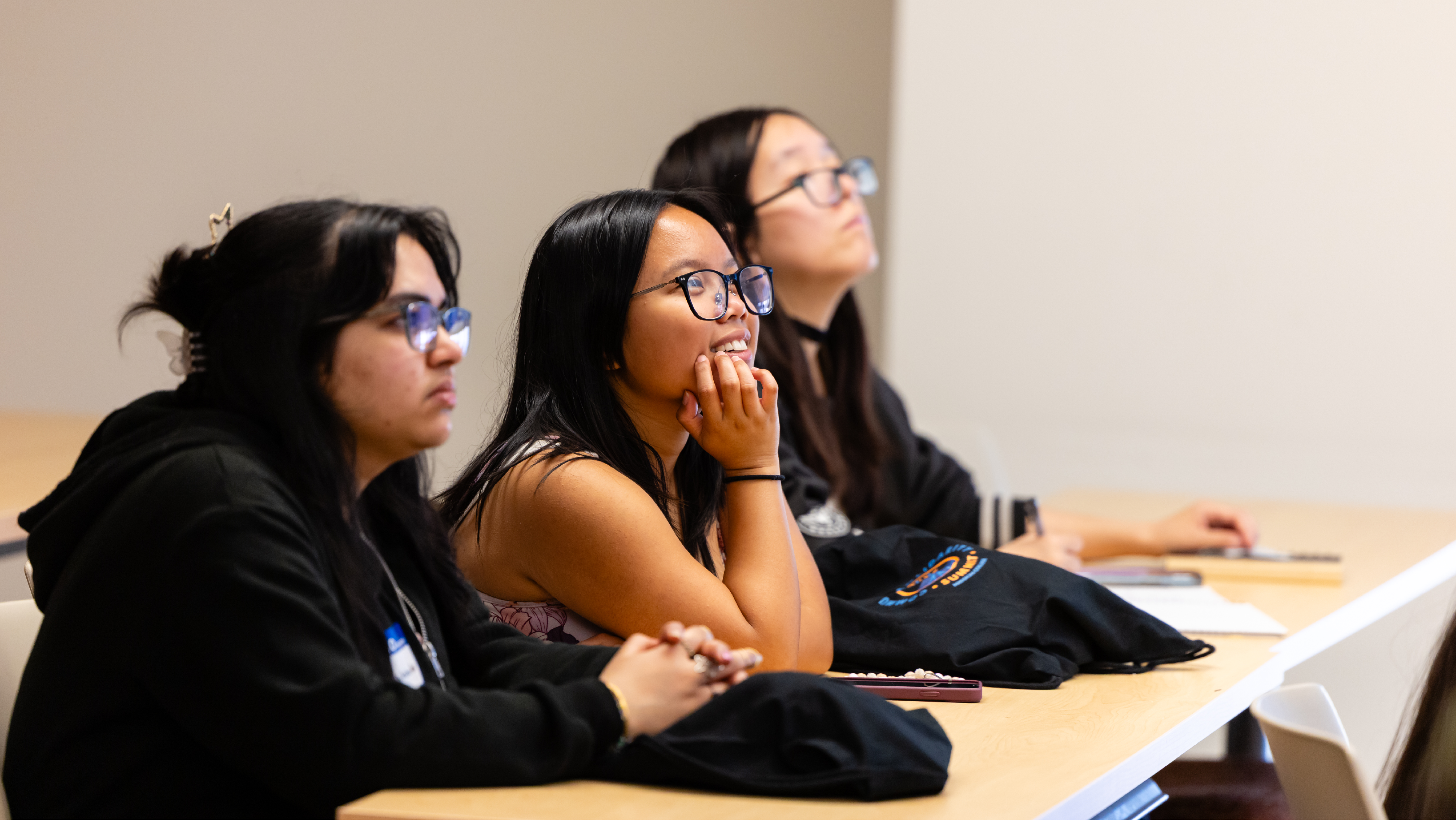 Three female students seated at a long desk in a classroom, listening attentively during a lecture.