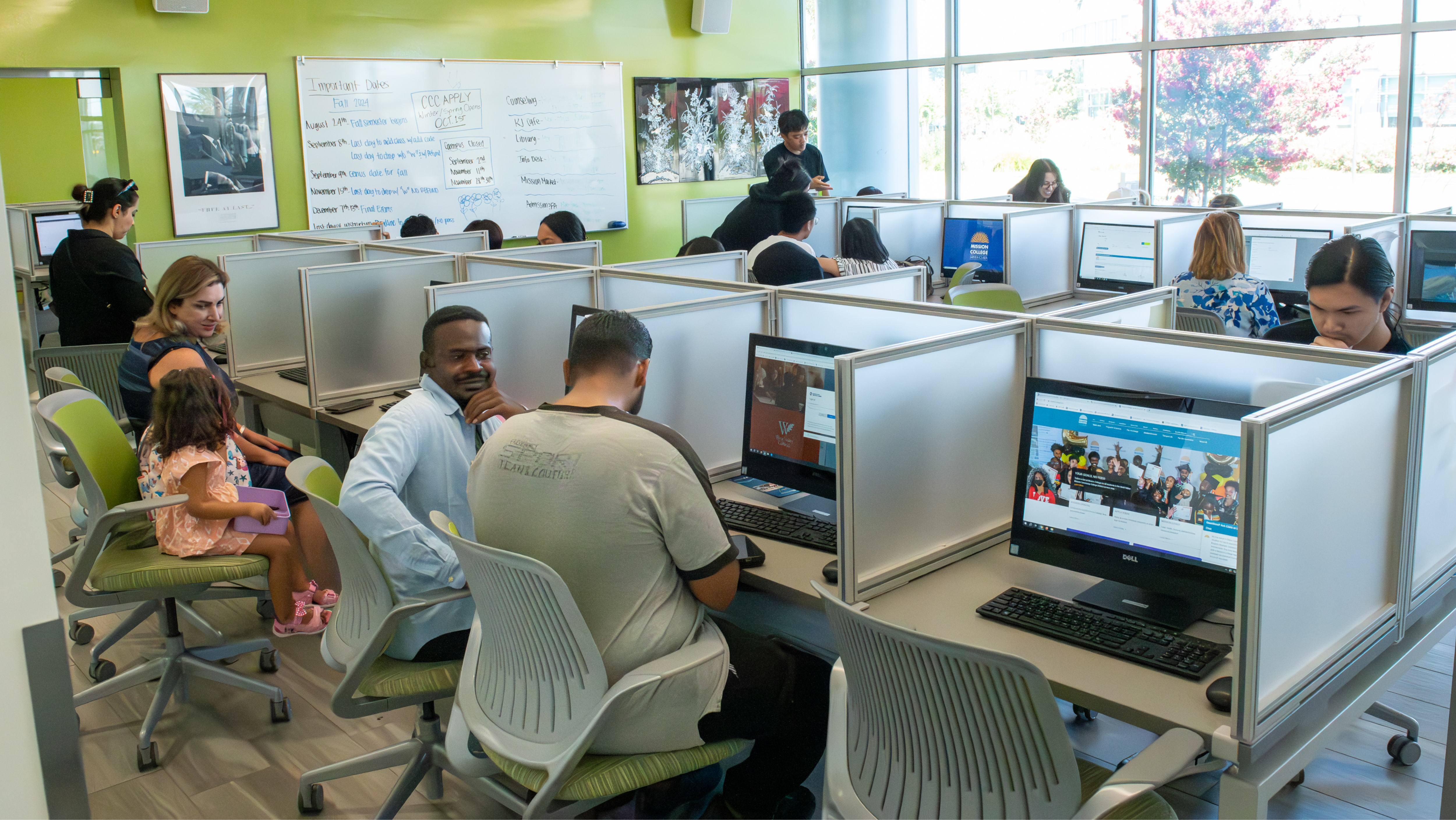 Students sitting at individual computer work cubicles in a brightly lit campus computer lab with a large whiteboard.