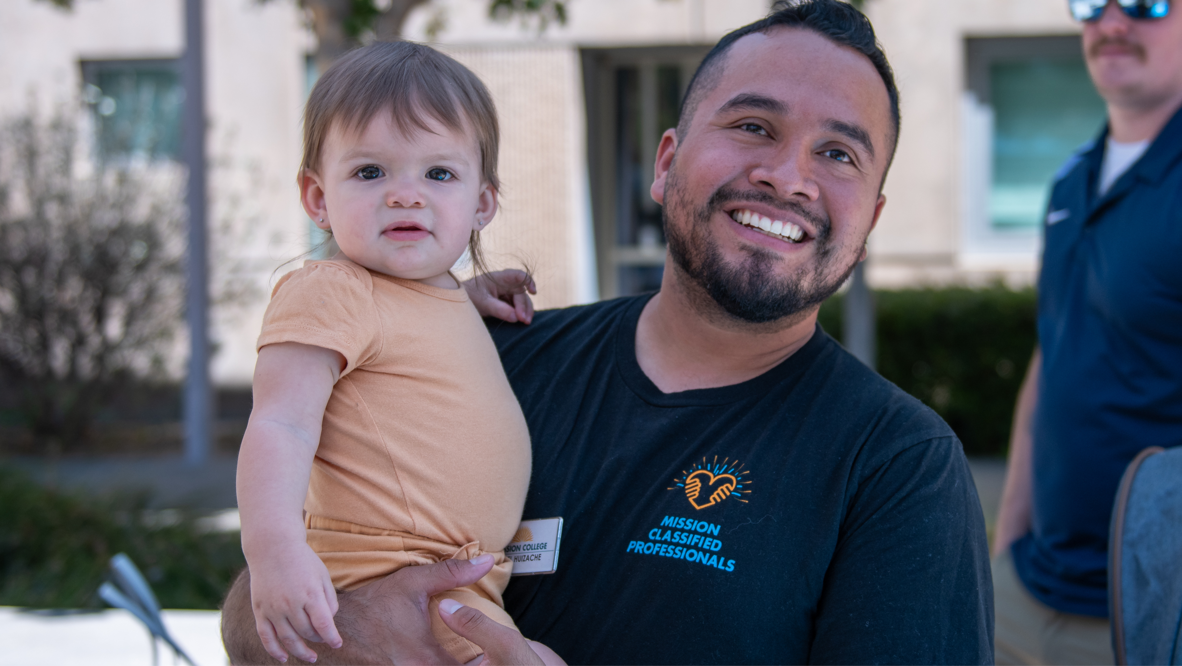 A smiling campus employee in a black shirt holding a young toddler outside during an event.