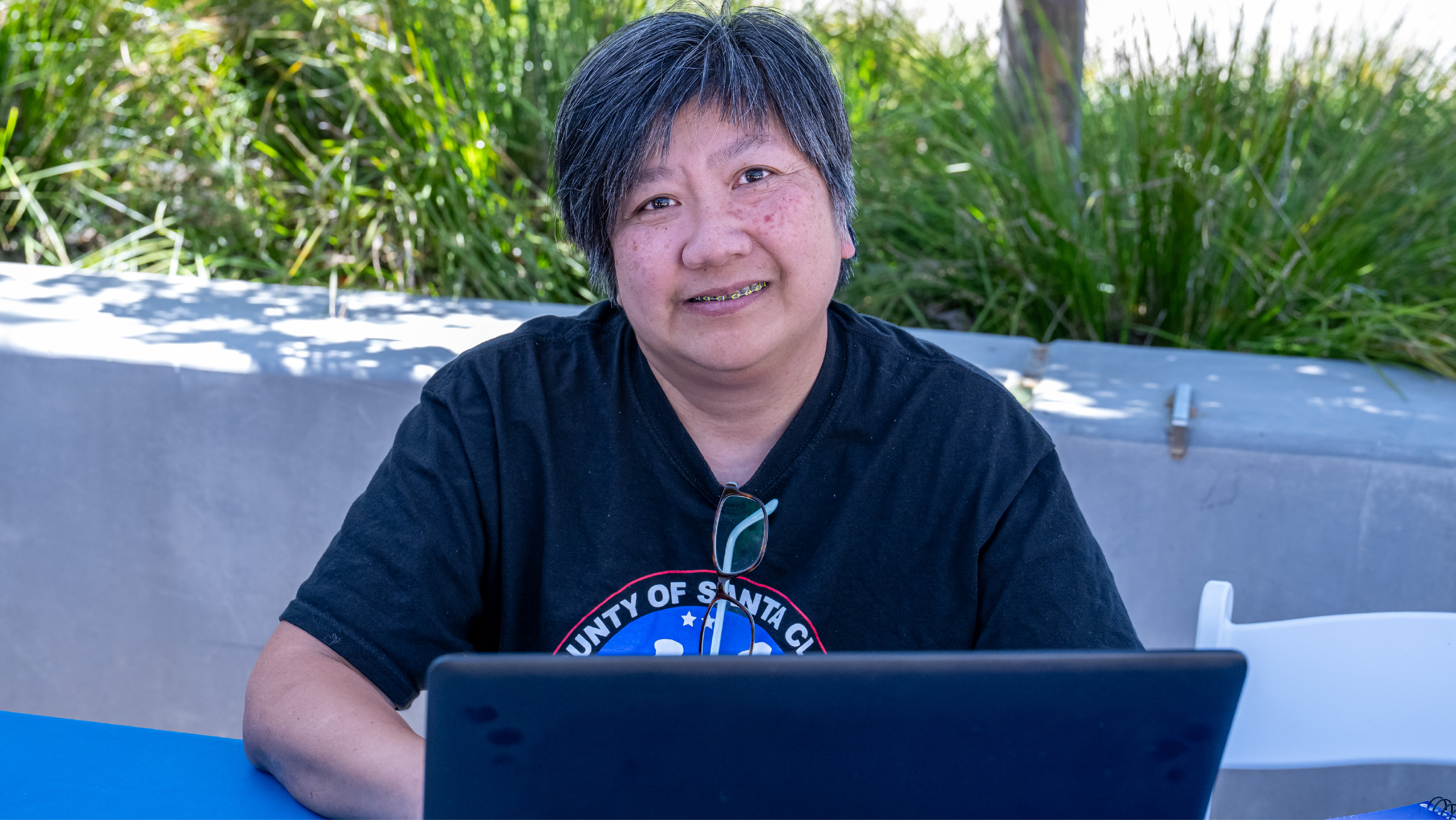 A person with short dark hair smiling while seated at a blue table in front of an open laptop.