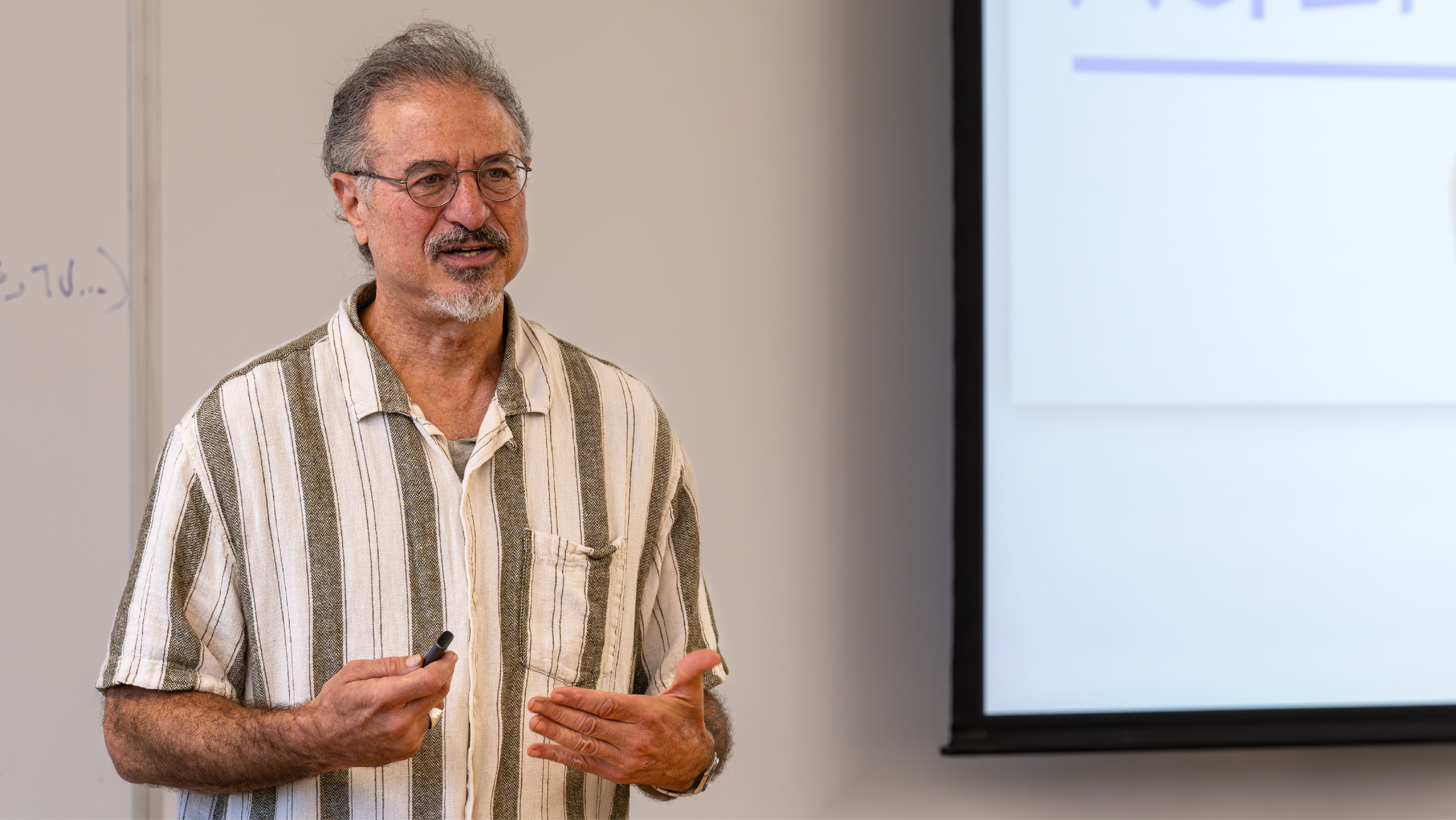 A male instructor with glasses and a striped shirt speaking during a presentation next to a projection screen.