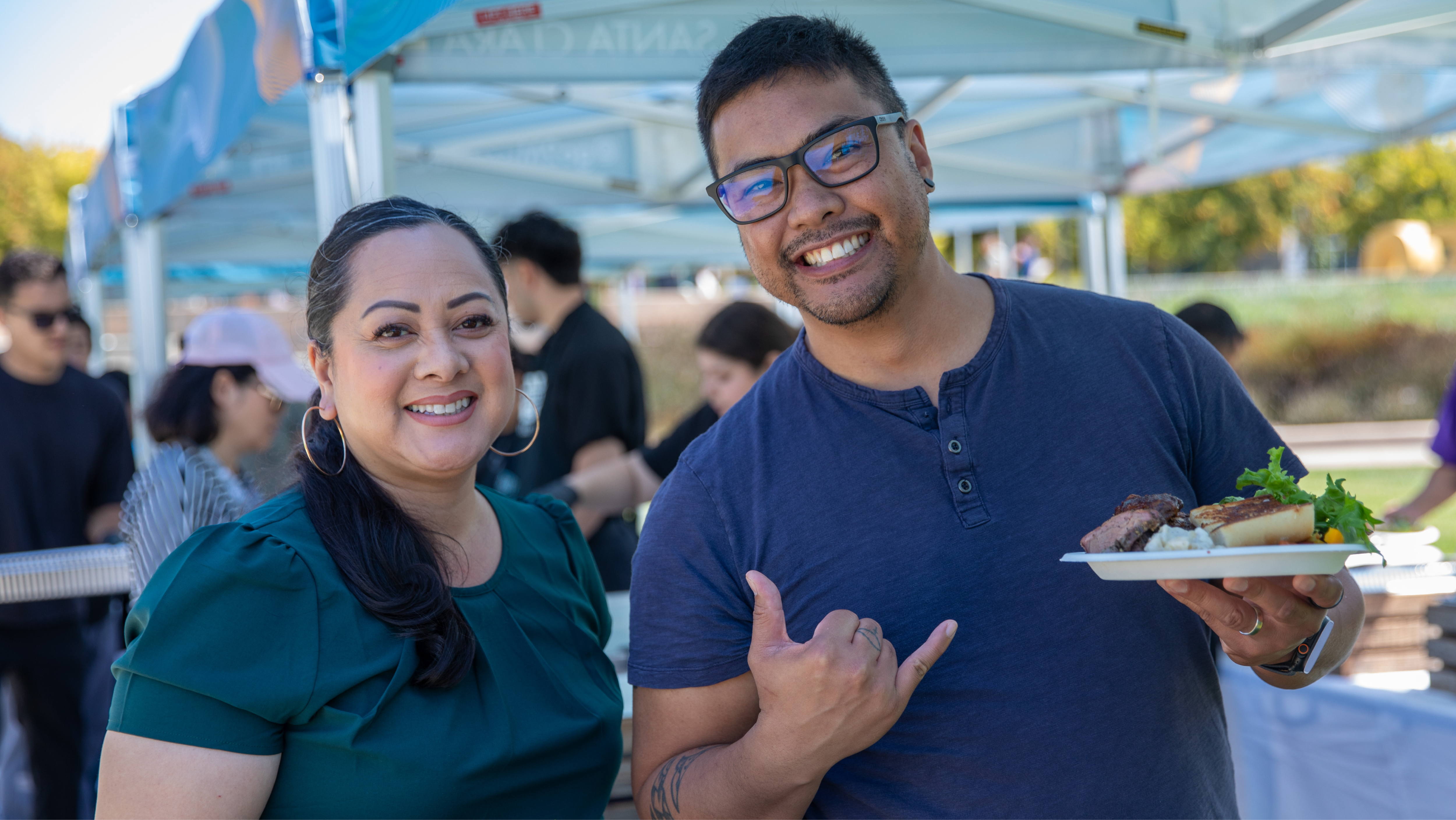 Two staff members smiling at an outdoor event, with one holding a plate of food and giving a shaka sign.