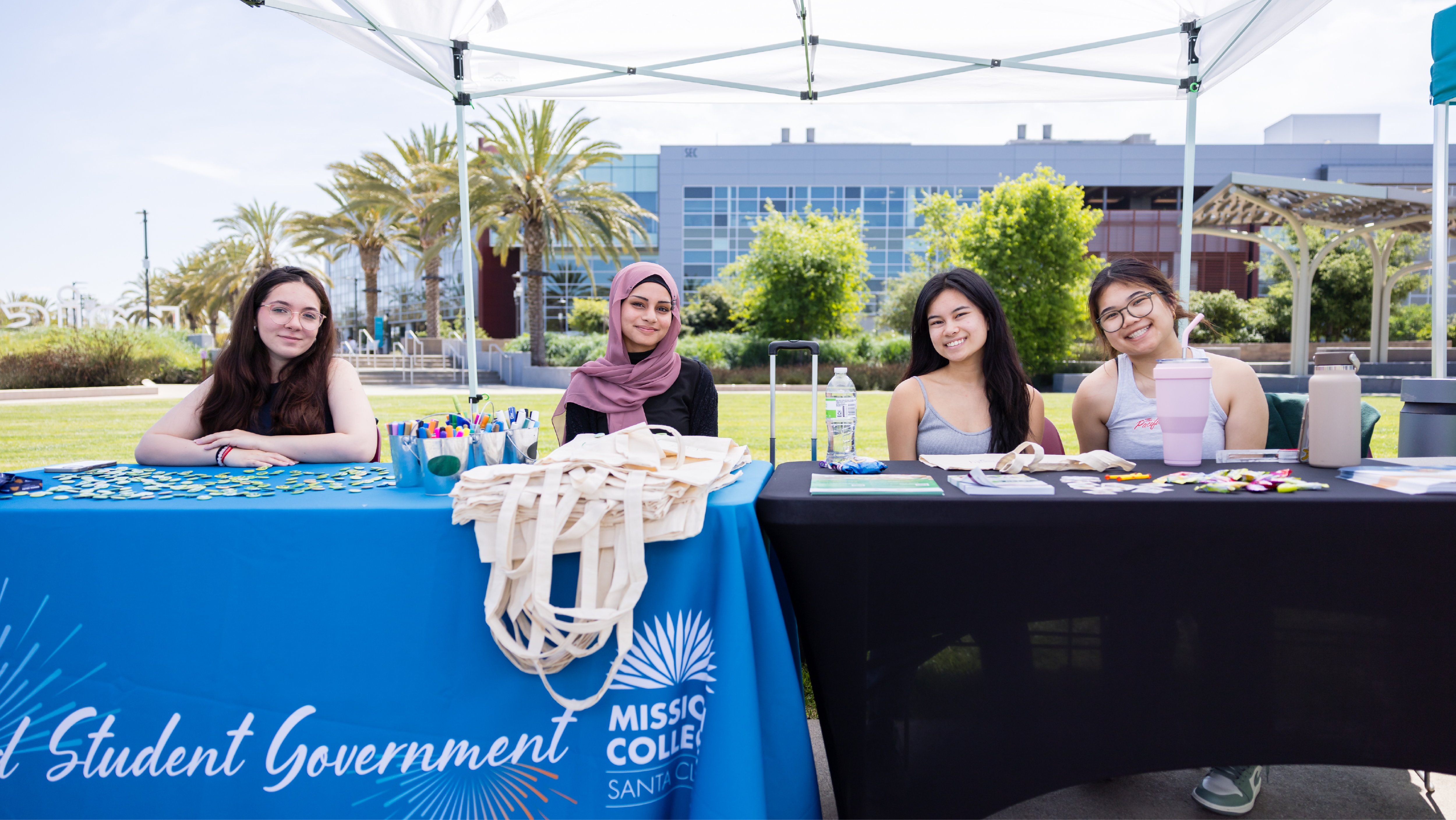 Four female students sitting behind an ASG canopy booth with promotional tote bags and supplies spread across the table.