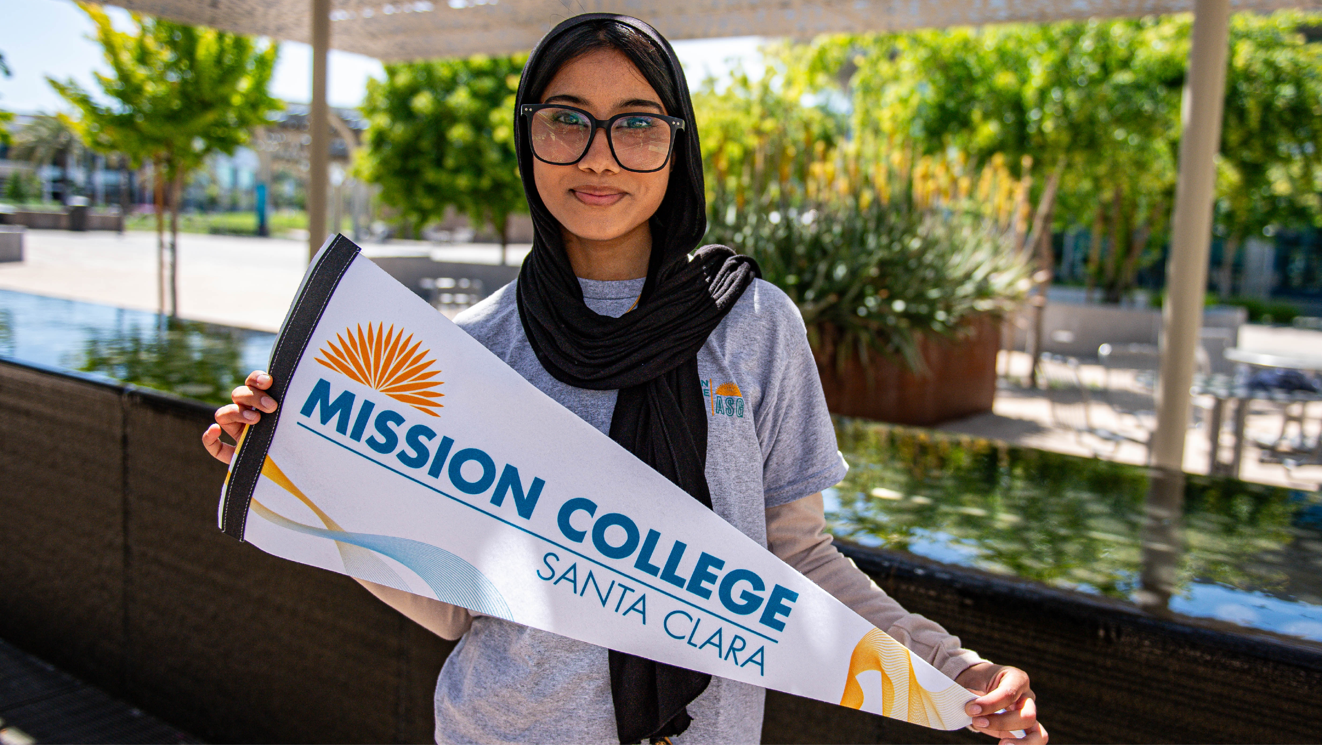 A student wearing a black hijab and glasses holding up a white and blue Mission College pennant flag outdoors.