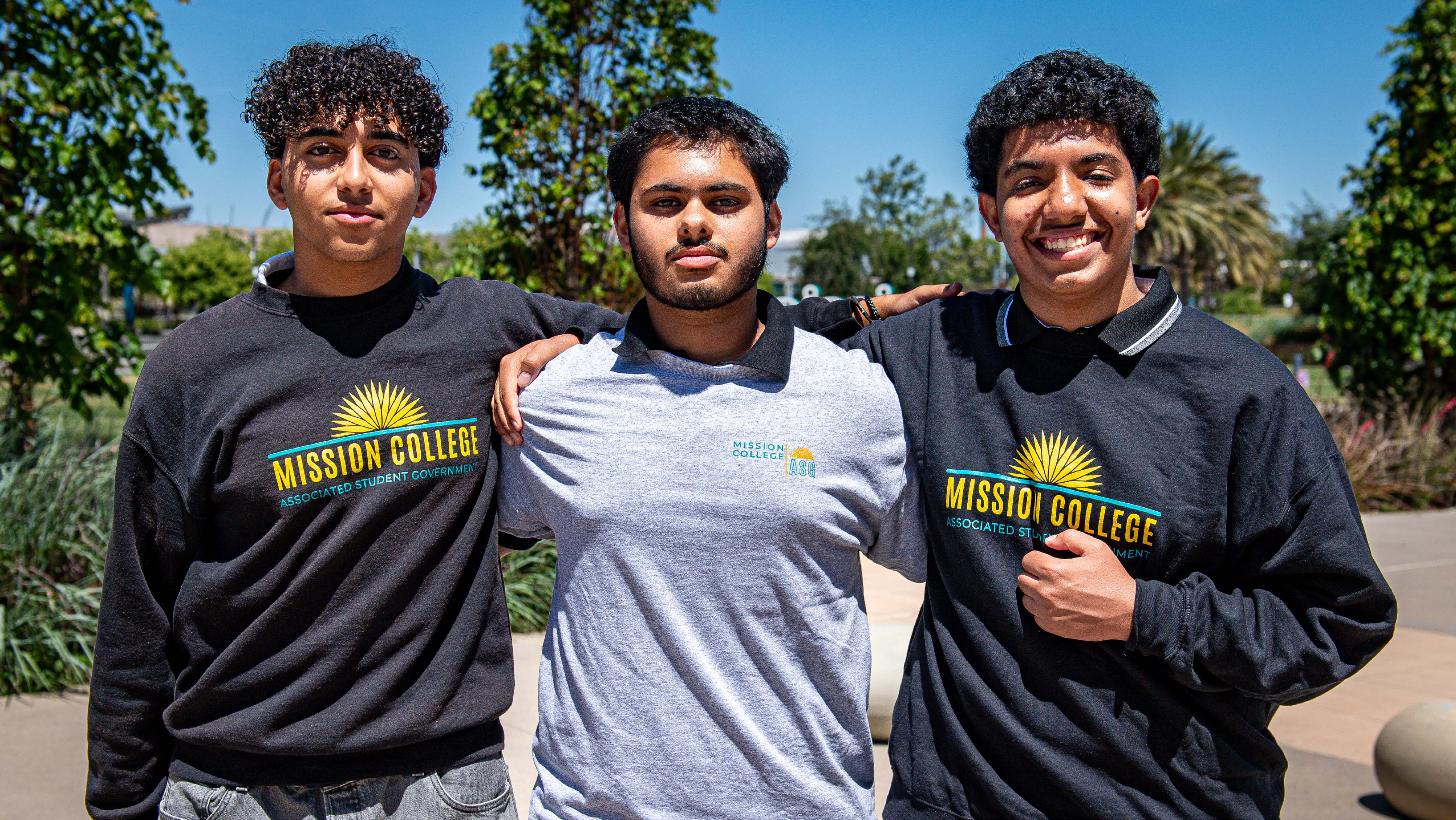 Three male students wearing Associated Student Government branded apparel posing arm-in-arm outdoors.