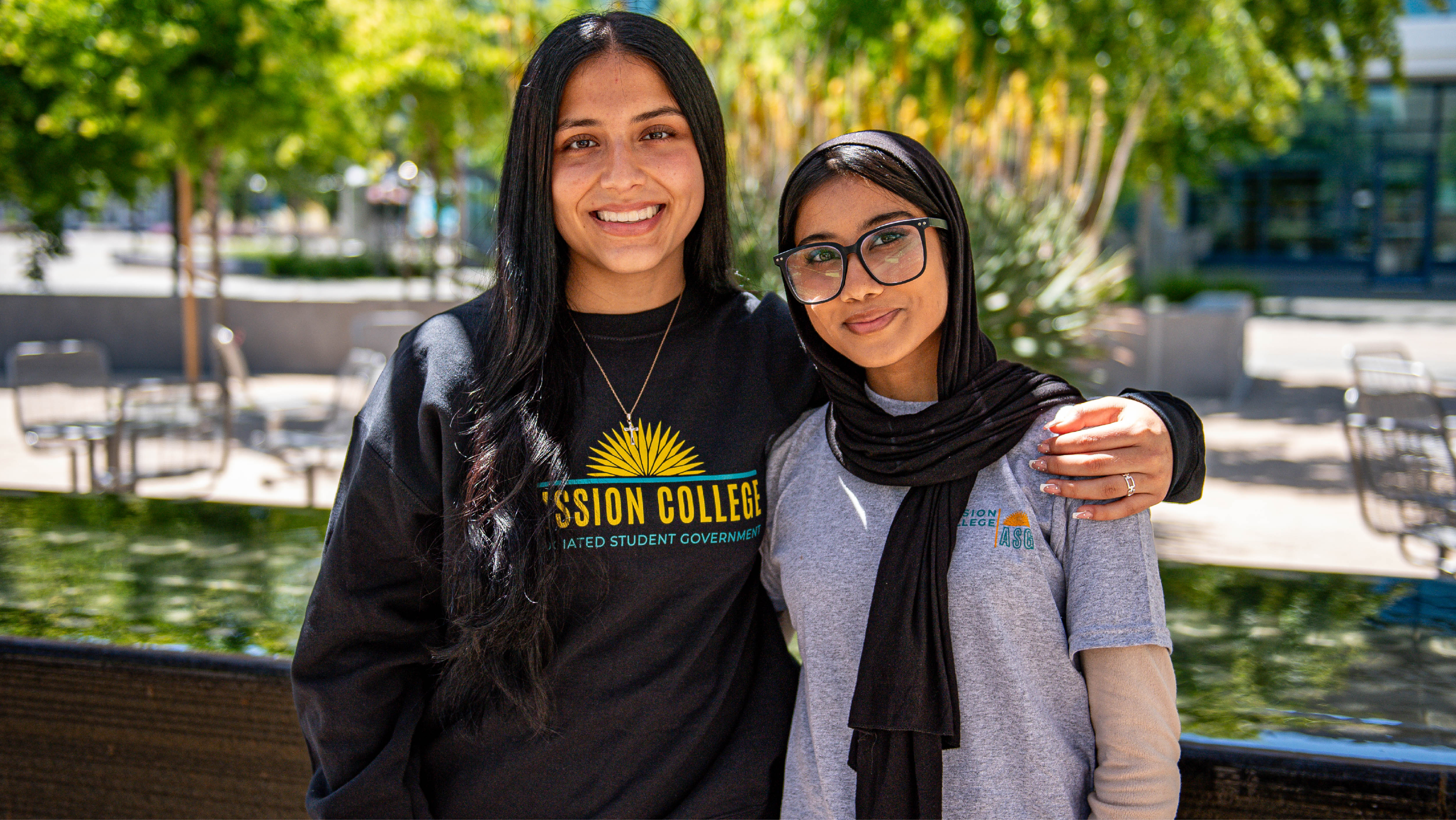 Two female ASG student leaders standing close and smiling together in front of a campus water feature.