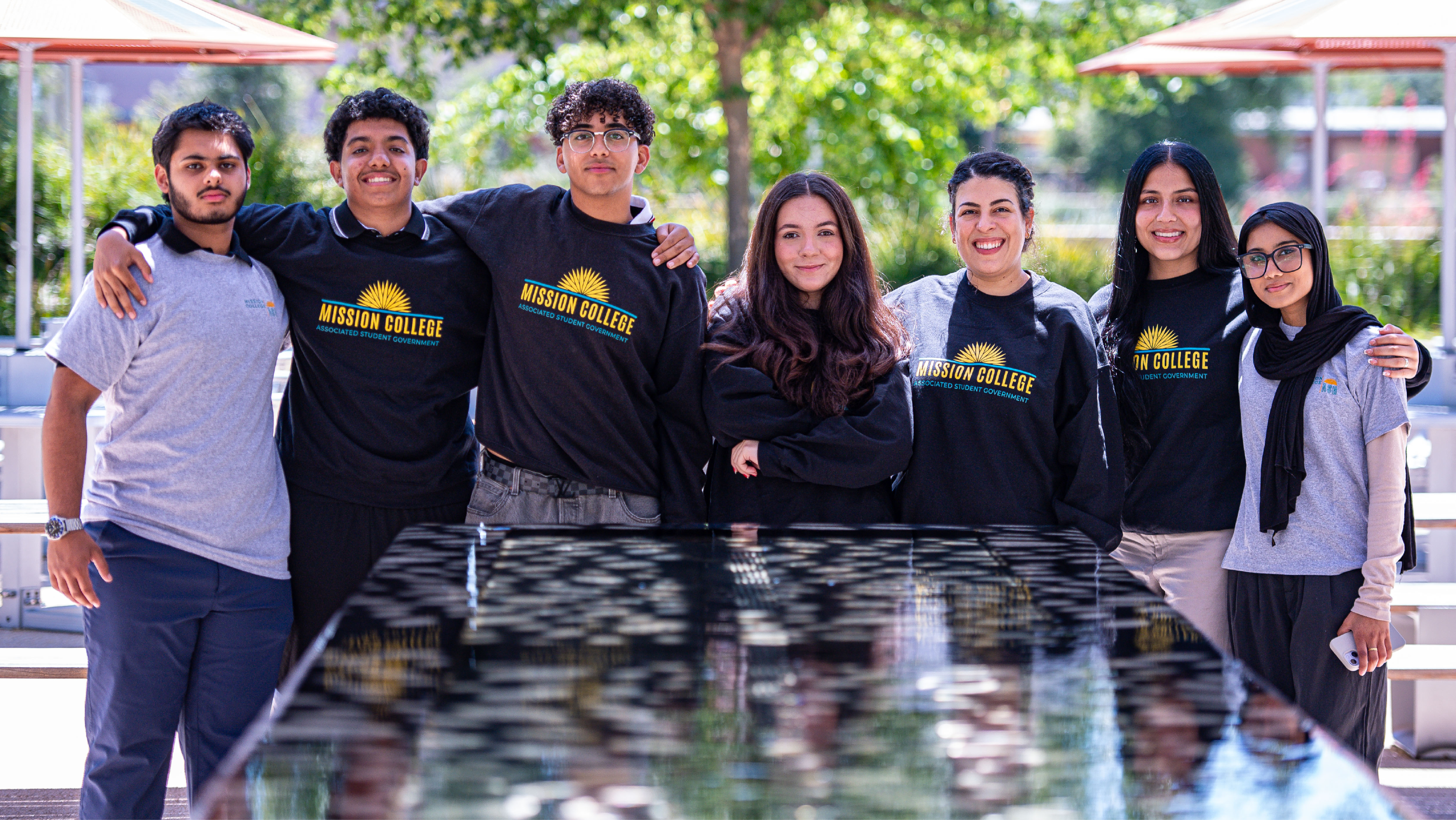 A group of seven ASG student officers posing behind a reflective dark table on a sunny outdoor patio.