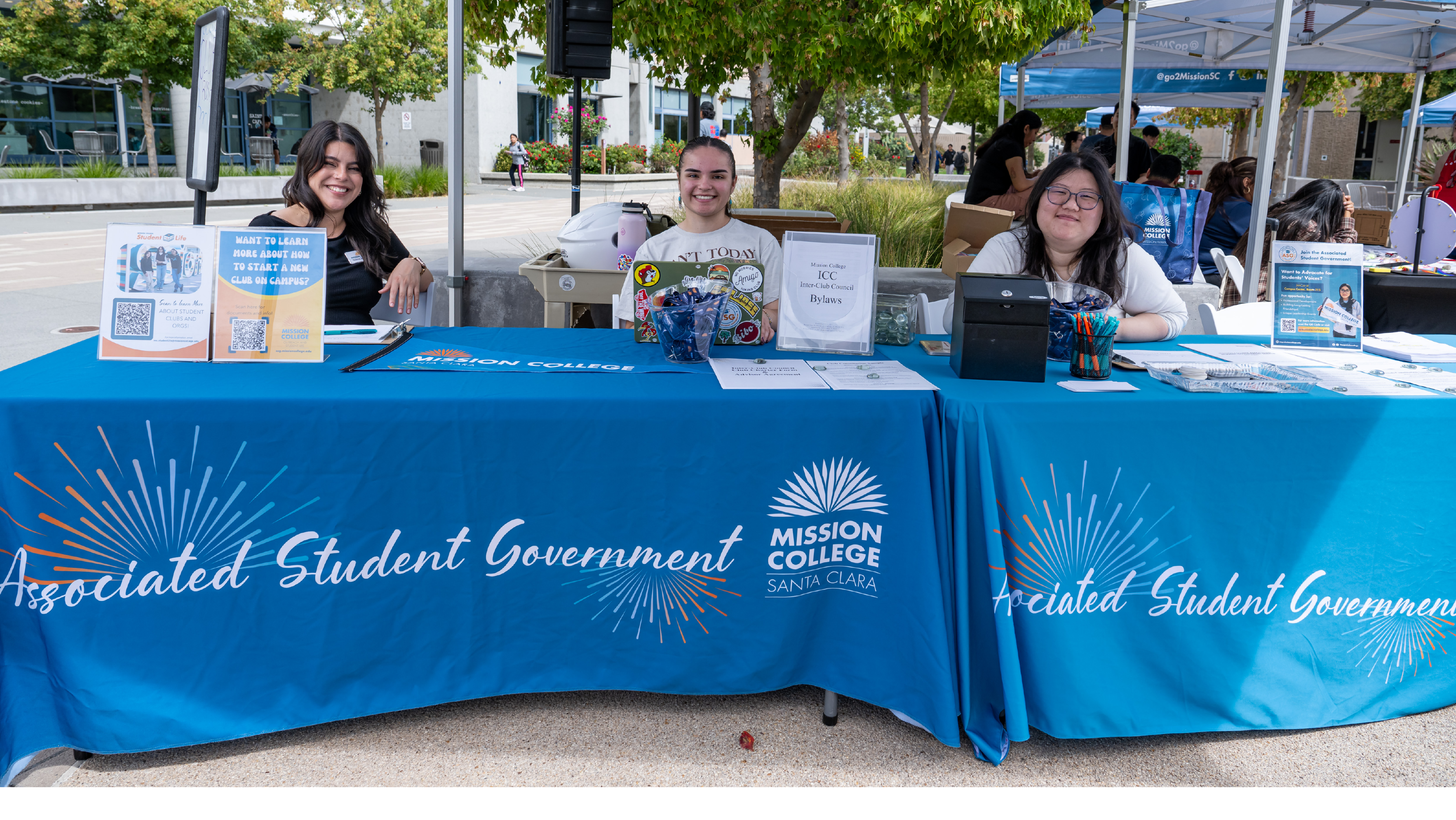 Three female student leaders sitting behind an Associated Student Government information table with flyers and promotional items.