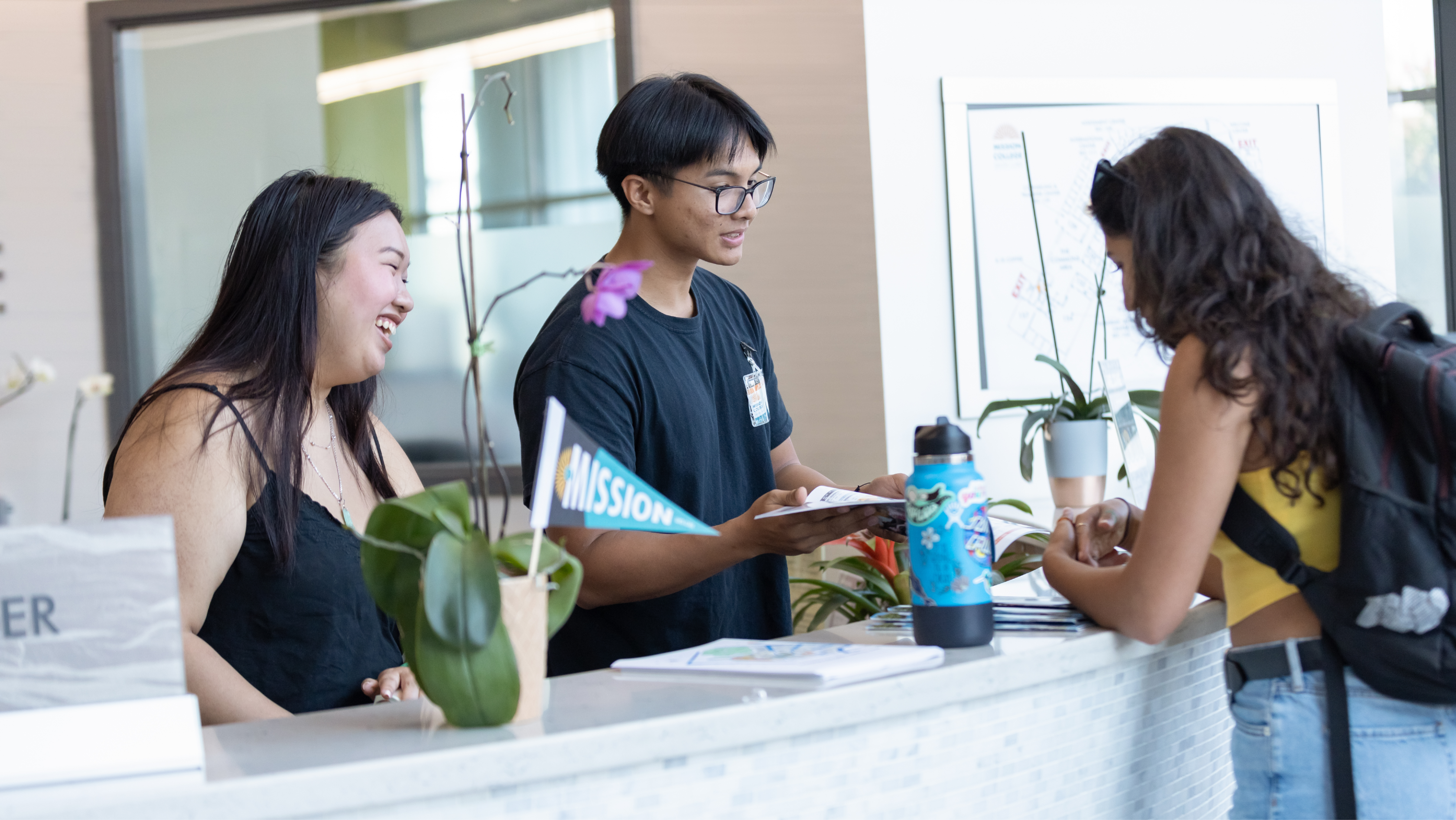 A student visiting an information desk counter while two staff members assist with printed schedules and pamphlets.