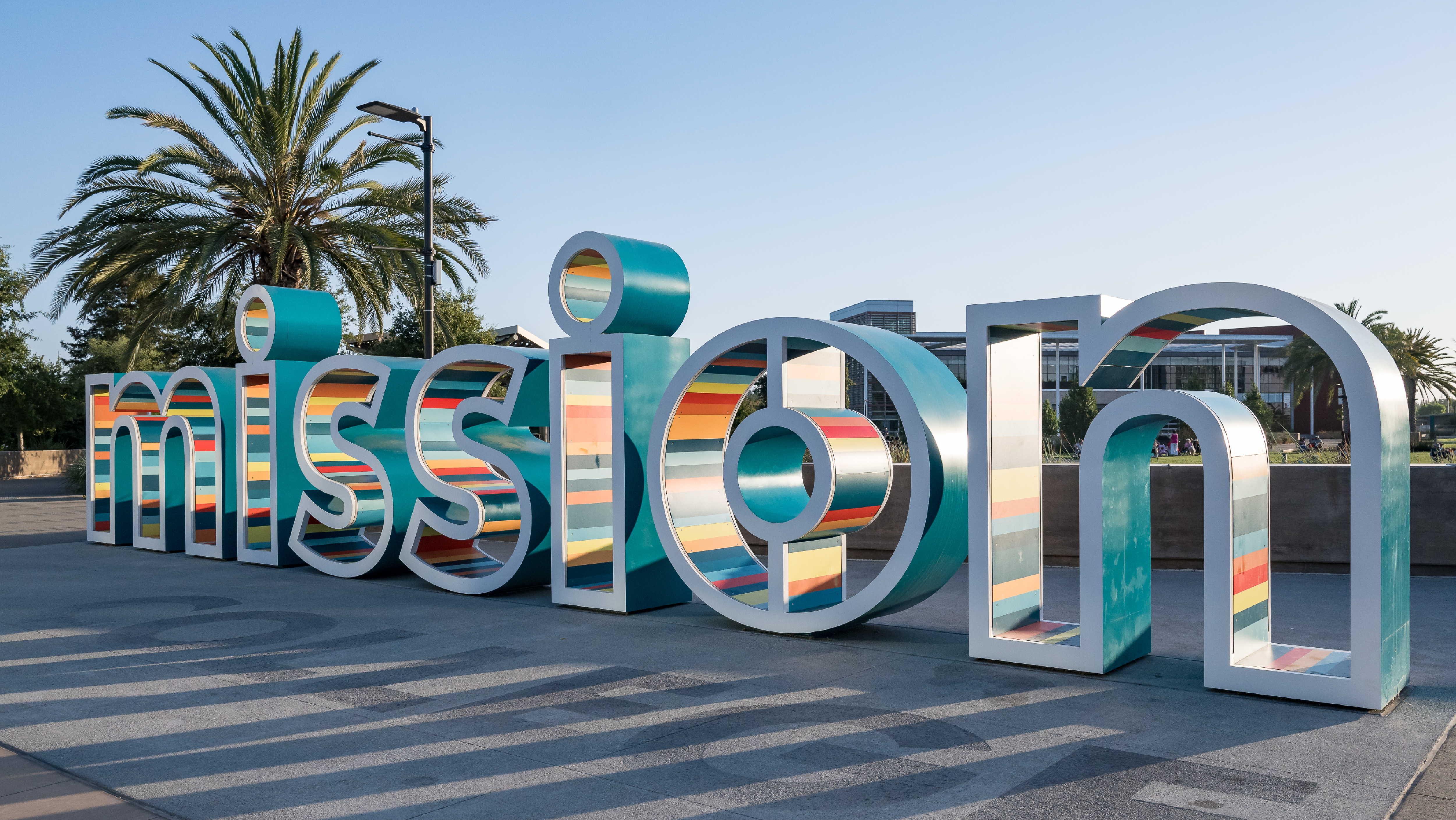 A wide view of the large, colorful "Mission" letter landmark sculpture on campus under a clear blue sky.