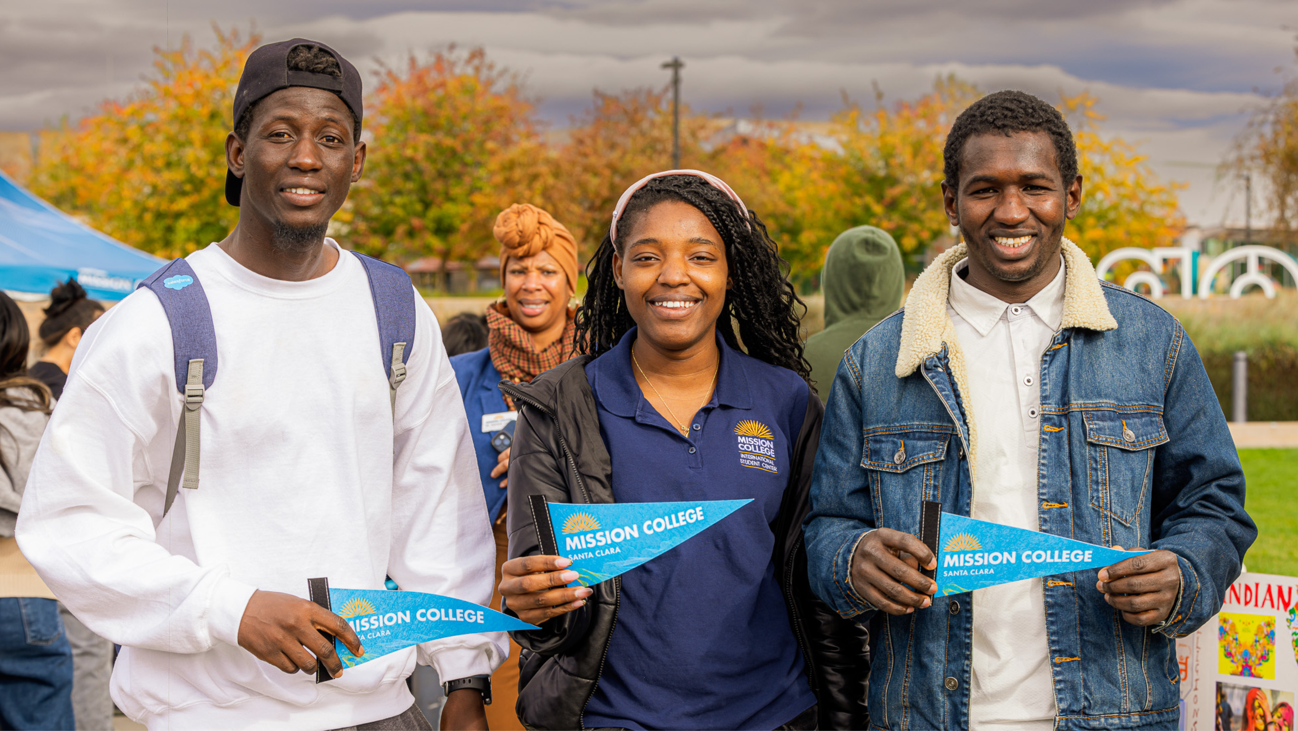 Three students standing outdoors holding blue Mission College pennant flags during an event.