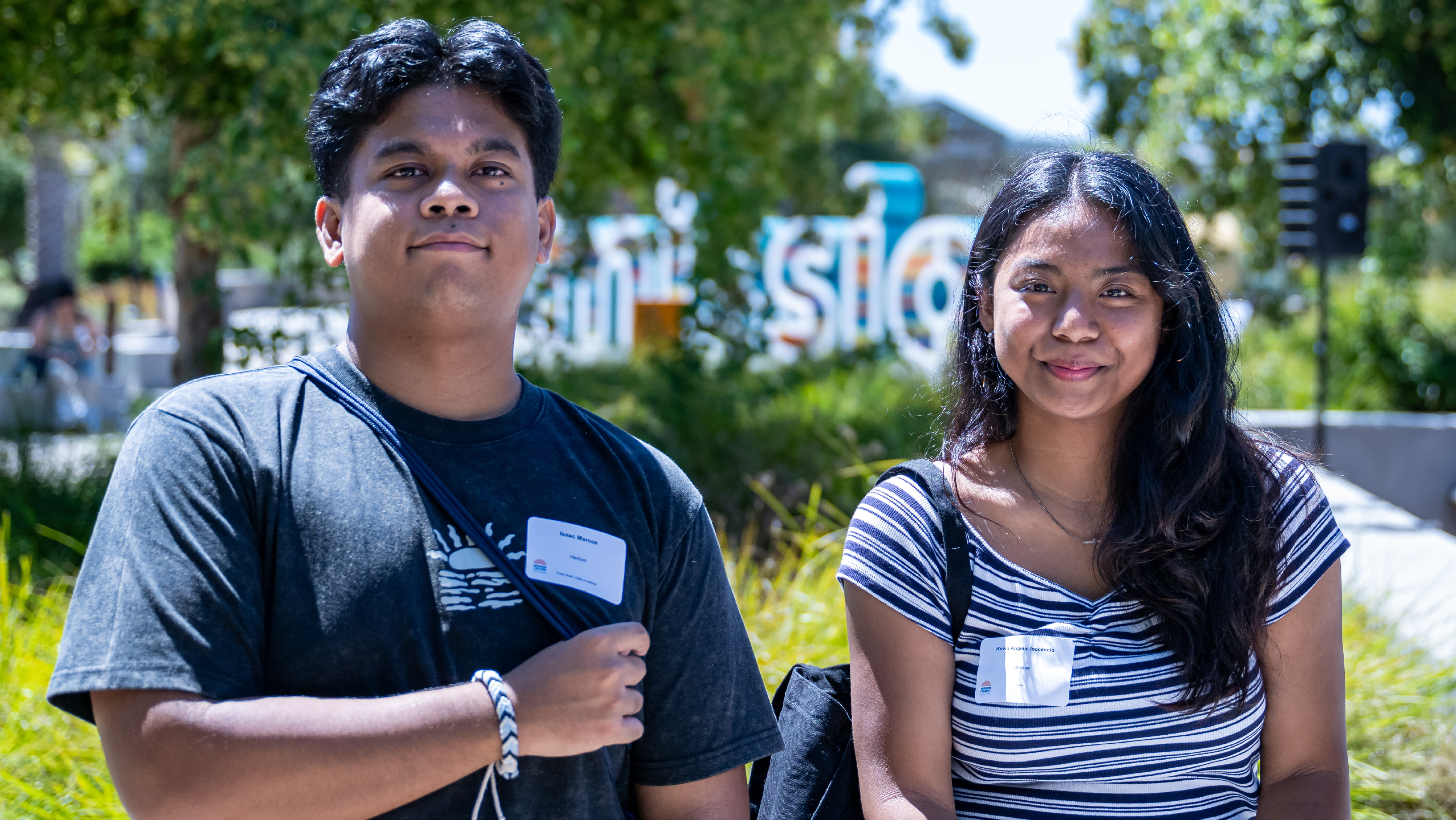 Two smiling students with name tags standing on campus in front of the Mission College letters.