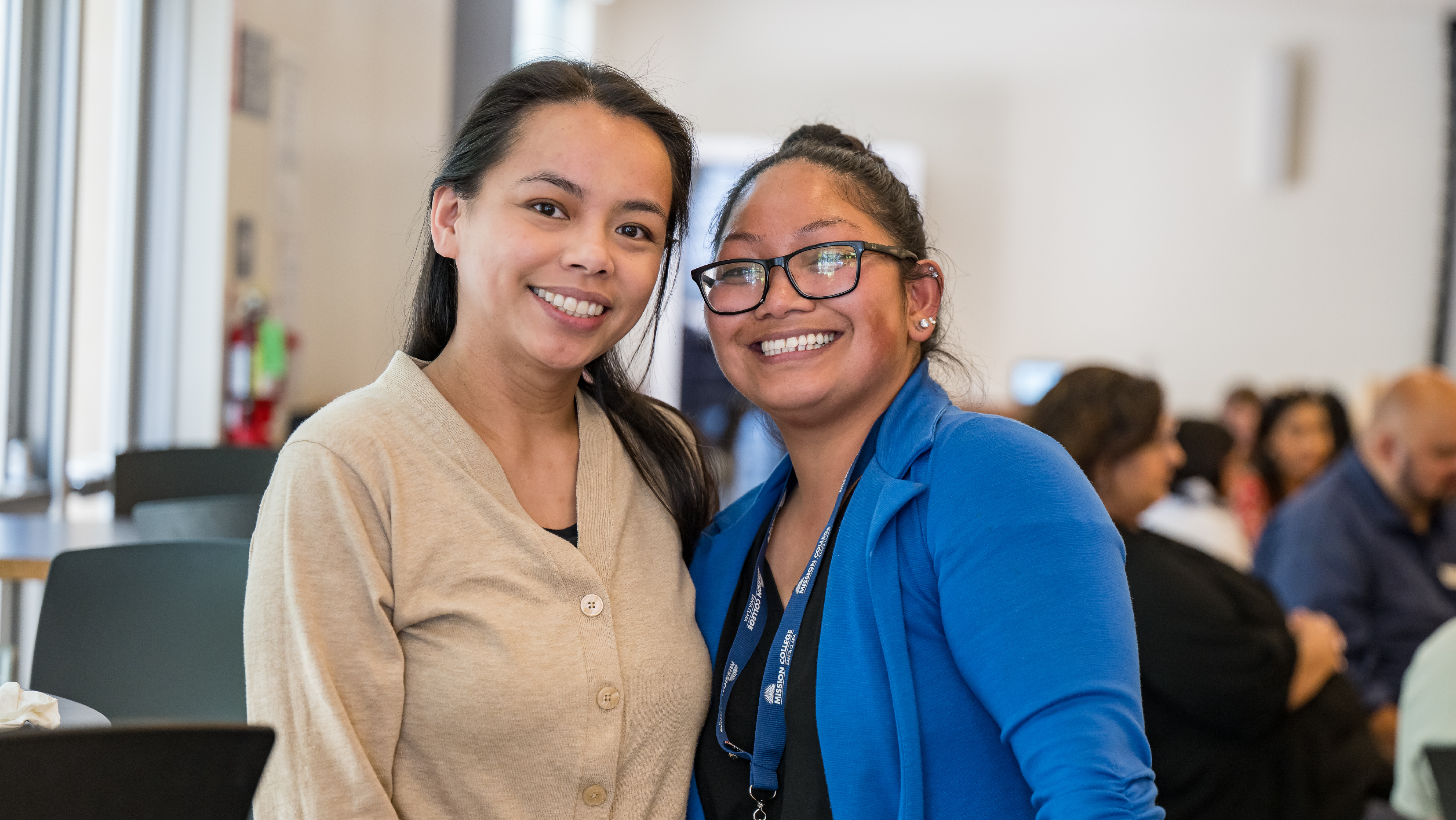 Two smiling students with name tags standing on campus in front of the Mission College letters.