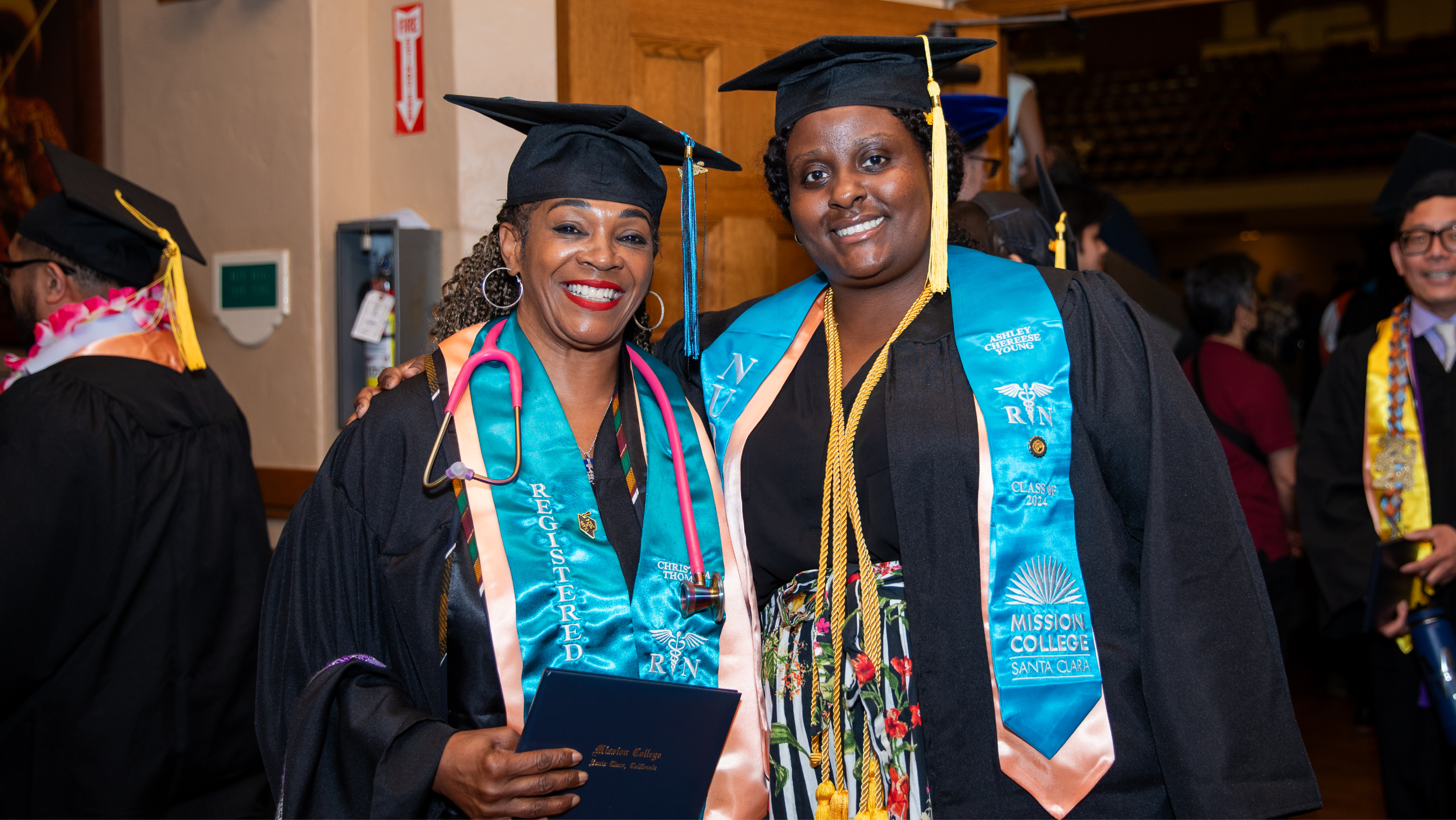 Five smiling Mission College graduates standing together in caps, gowns, and ceremonial stoles.