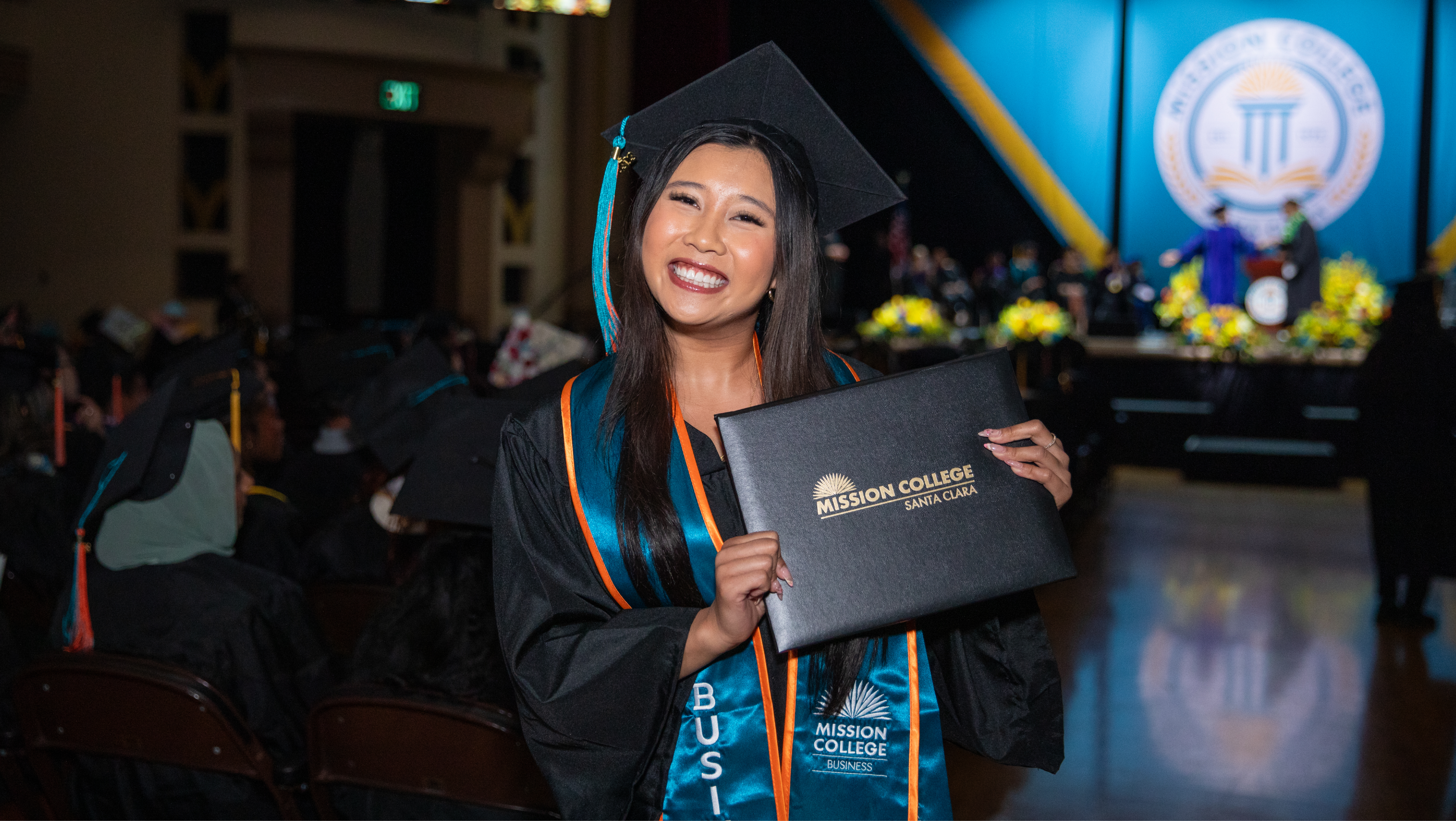 Mission College graduate proudly holding her diploma cover above her head on stage in front of the college logo backdrop.