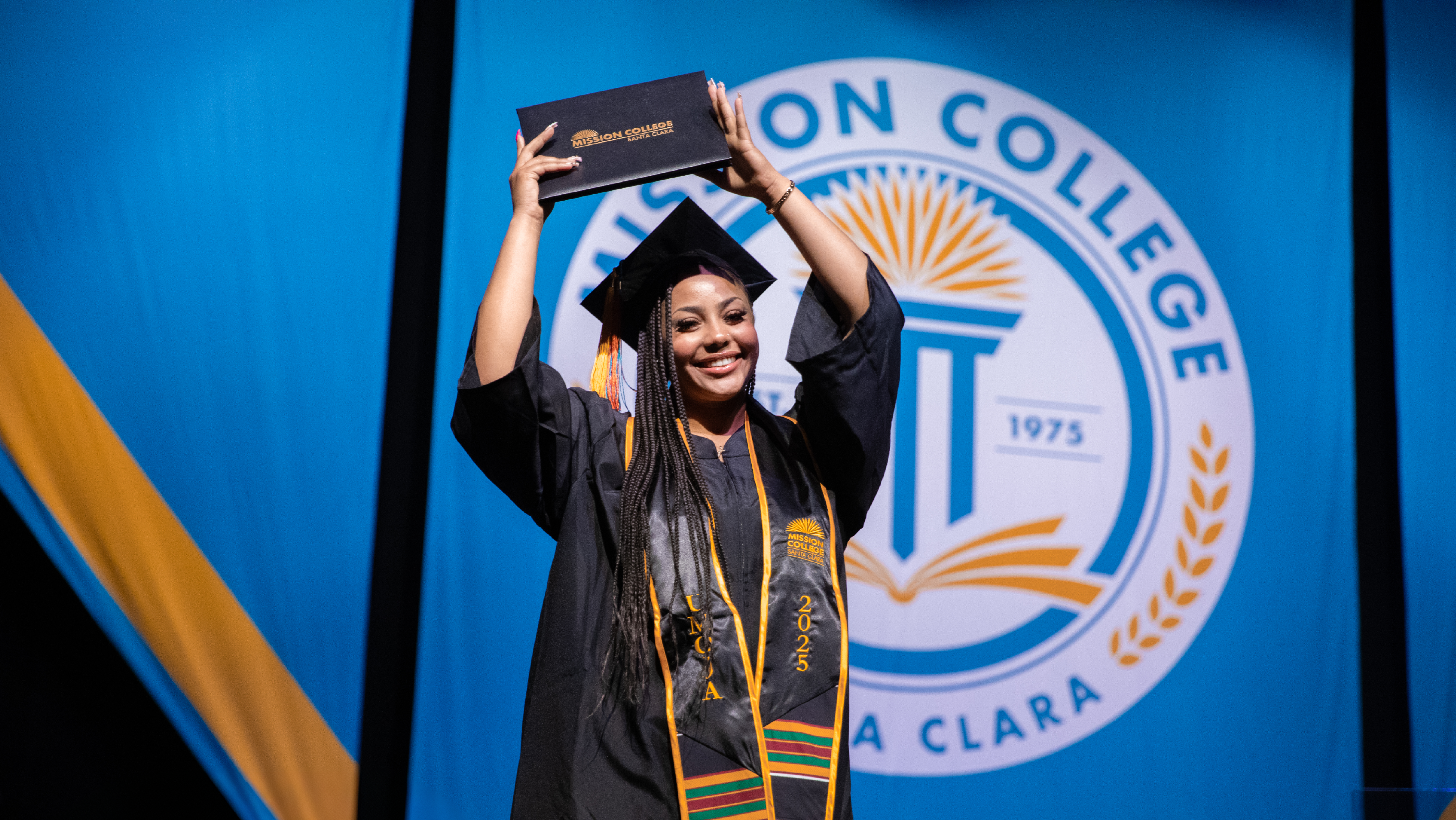 Mission College graduate in cap and gown waving happily at San Jose Civic venue.