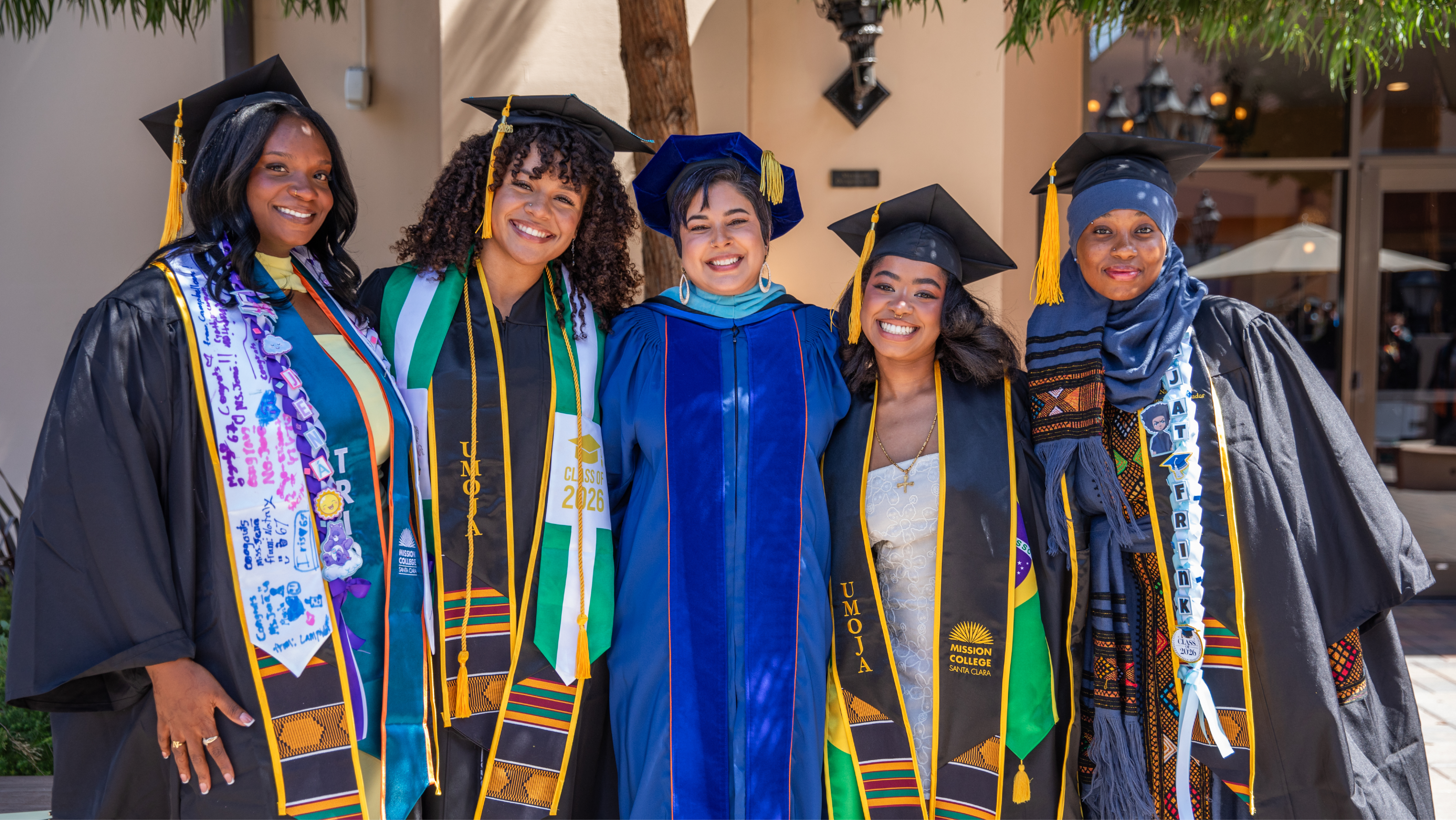 Graduate wearing leis and holding flower bouquets celebrating outdoors with laughing friends and family.