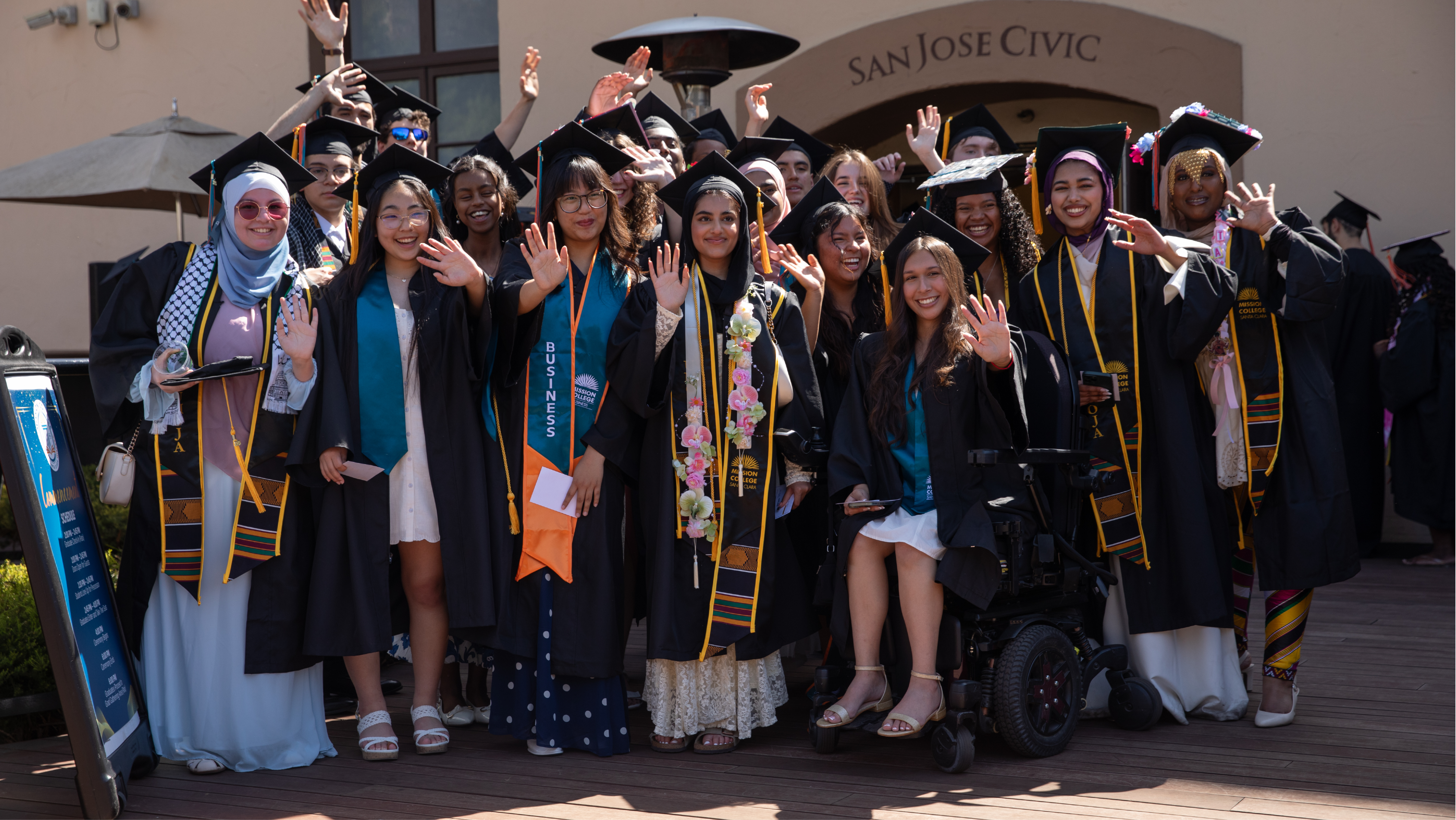 Group of Mission College graduates posing together in caps, gowns, and teal sashes holding diploma covers.