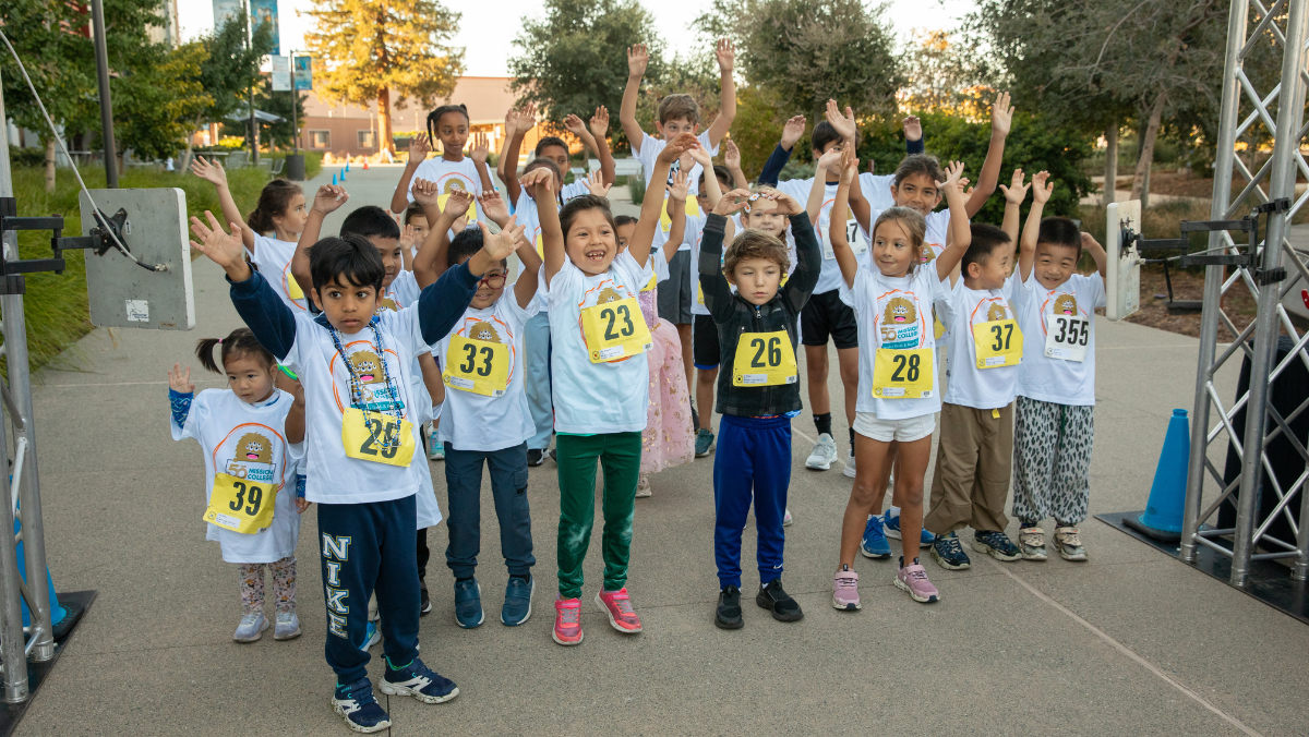 Children wearing race bibs and Monster Dash & Bash T-shirts raise their hands excitedly at the Kids Fun Run starting line on the Mission College campus.