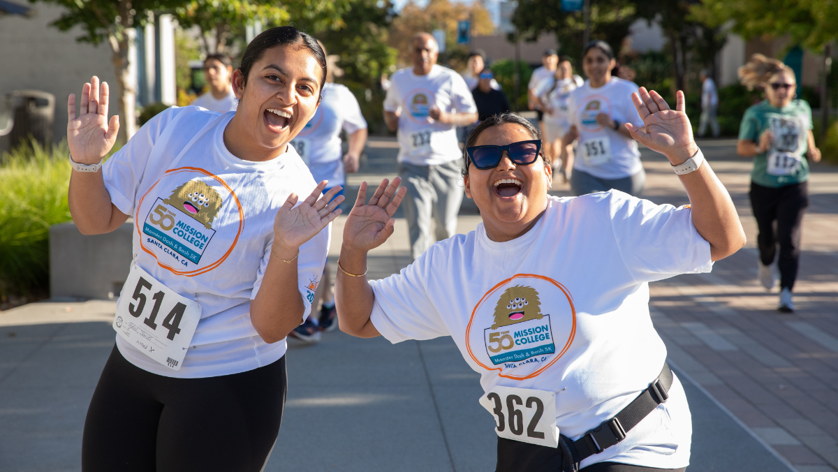 Two participants smile and wave to the camera while walking the Monster Dash & Bash 5K.