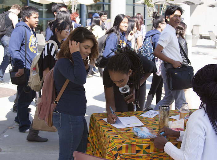 Girl leans over table to get information about a club on Club Day. Student mill around in background.