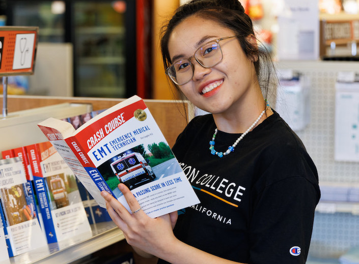 Young woman of Asian descent wears a black Mission College tshirt and holds an EMT book in the Bookstore. She wears metal glasses.
