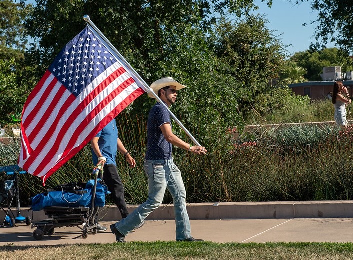 Young Latino man in cowboy hat and jeans carries large American flag.