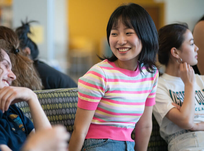 Female college student of Asian descent is socializing with a group of students. She is wearing a pink and white striped shirt and has shoulder-length black hair.