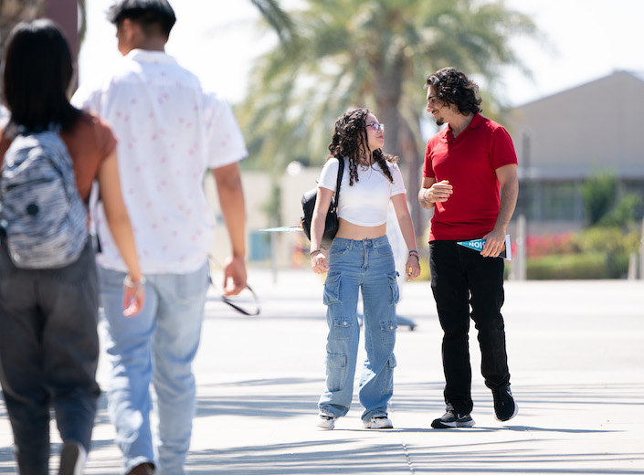 Young woman with curly dark hair and short white tshirt walks with young man in red tshirt and jeans on college campus.