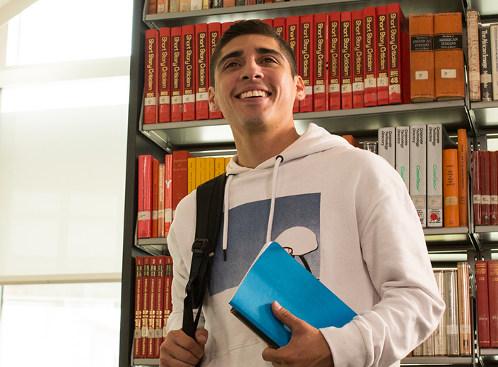 Latino college student holds books in this right-hand in the library. He wears a white hoodie and smiles.