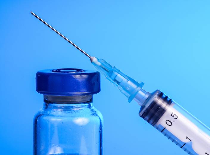 A needle and small clear glass bottle of vaccine are pictured against a deep blue background.