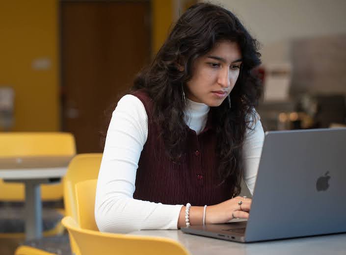 Young woman in red vest over long-sleeved shirt works on a laptop.