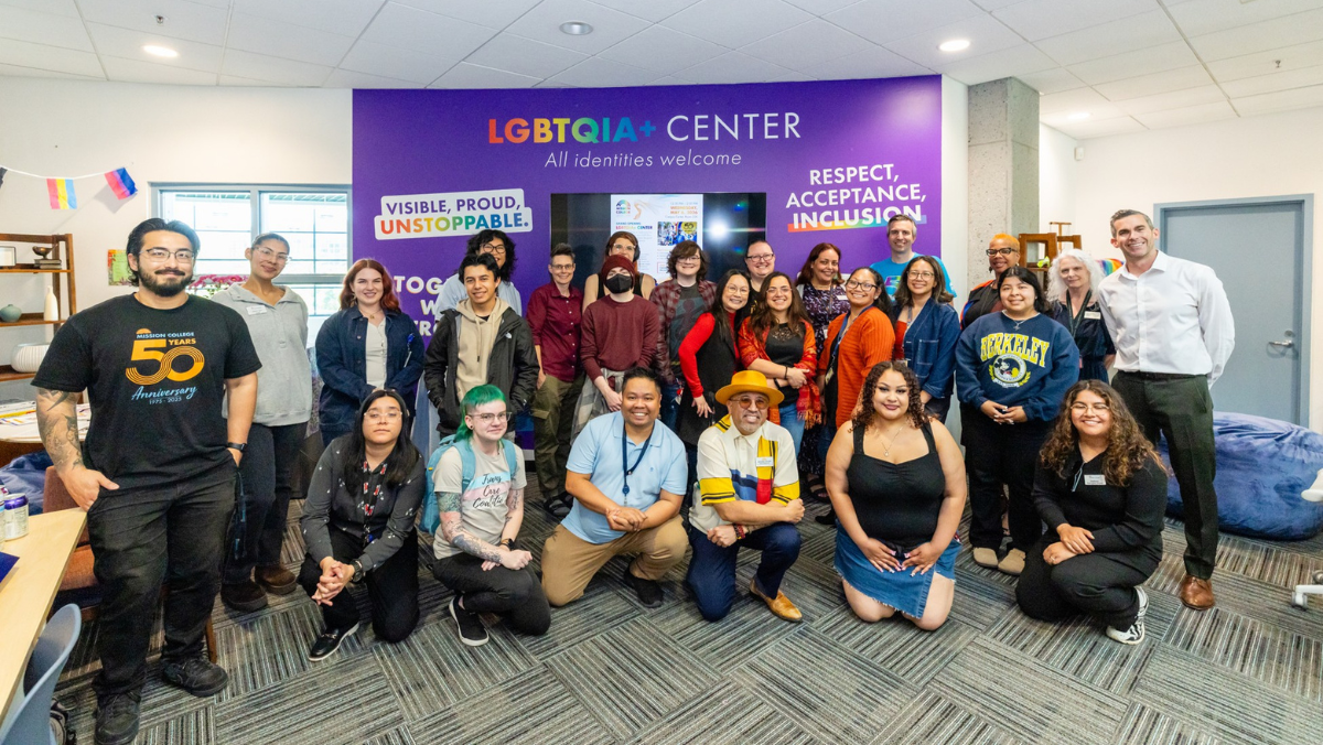 A diverse group of people gathering for a photo inside the LGBTQIA+ Center in front of a purple wall that reads "LGBTQIA+ Center - All identities welcome."