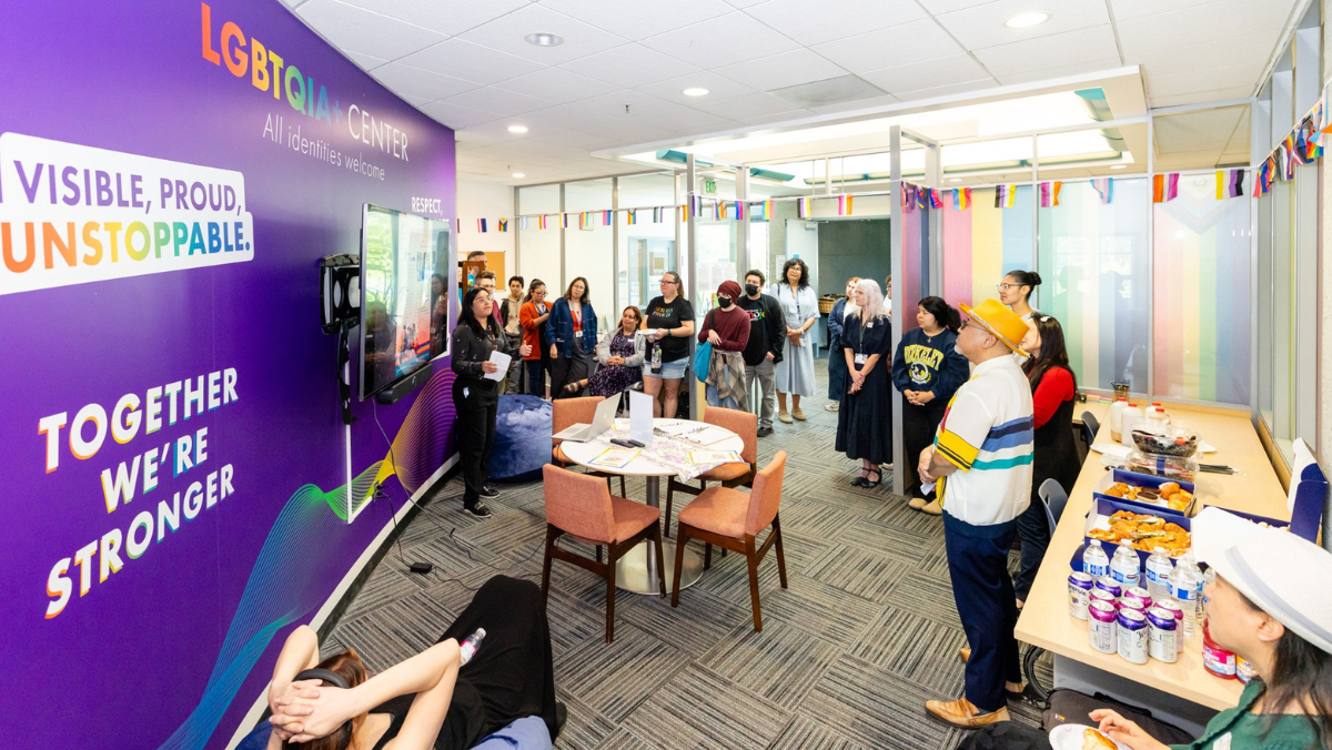 A diverse group of people gathering for a photo inside the LGBTQIA+ Center in front of a purple wall that reads "LGBTQIA+ Center - All identities welcome."