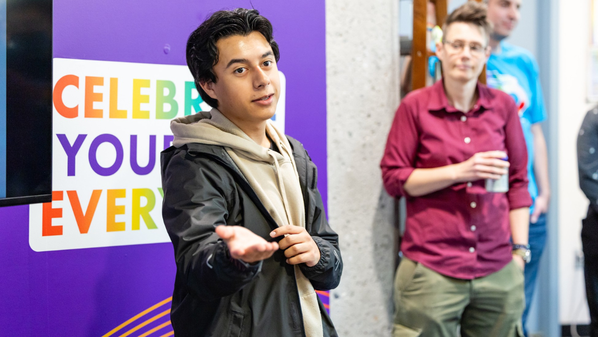 A young person wearing a black jacket and beige hoodie smiling and gesturing forward with an open hand during a presentation in front of a purple LGBTQIA+ Center sign.