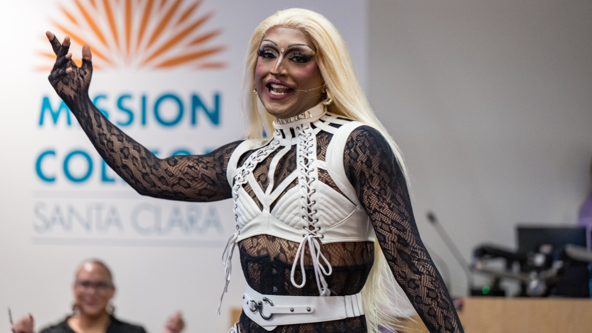 A person with long blonde hair and a detailed white and black laced outfit poses energetically indoors with a Mission College logo behind them.