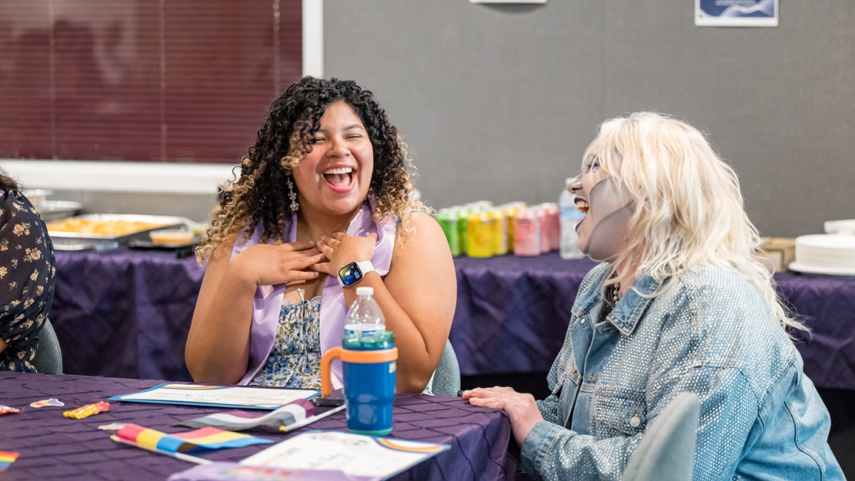 Two people sitting together at a purple-covered table with Pride flags, laughing joyfully during an event. One person wearing a purple stole holds their hands to their chest in delight while looking at their companion in a denim jacket and silver makeup.