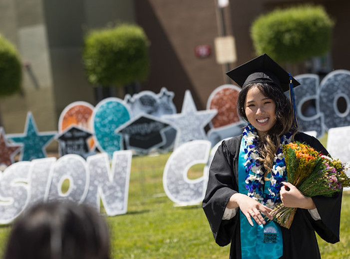 Young woman of Asian descent holds a bouquet in her cap and gown in from of Mission College signs on the Central Plaza.