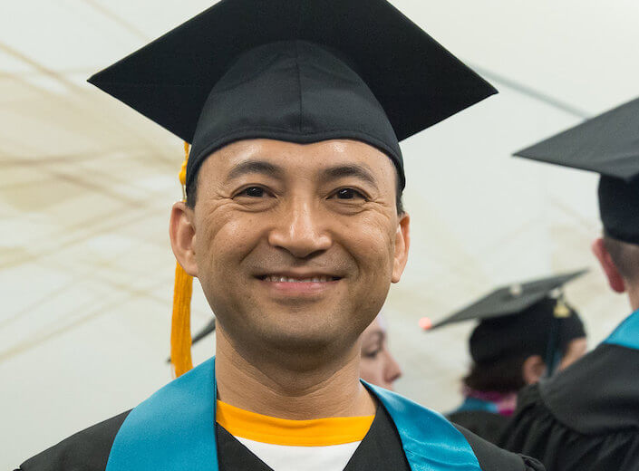 Adult/middle-aged man of South Asian or Hispanic descent wears a cap and gown with a yellow tassle. 