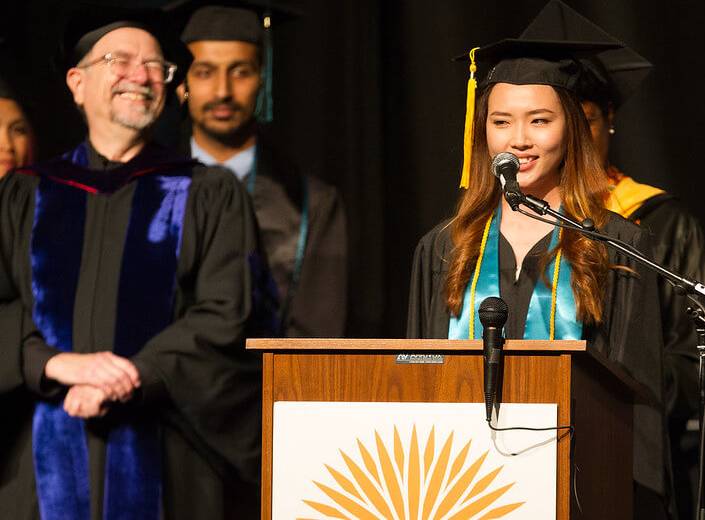 A young woman with long dark hair and graduation regalia on speaks at a podium. Behind her are other people in their commencement clothesm, and to the left of her is an older white faculty member in regalia laughing.