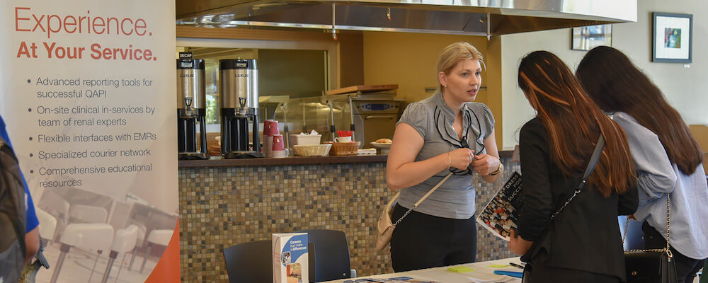 A woman with blonde hair stands at a table a recruiting event speaking to a student. 