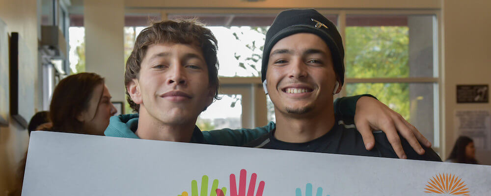 Two young man pose together holding a sign. 